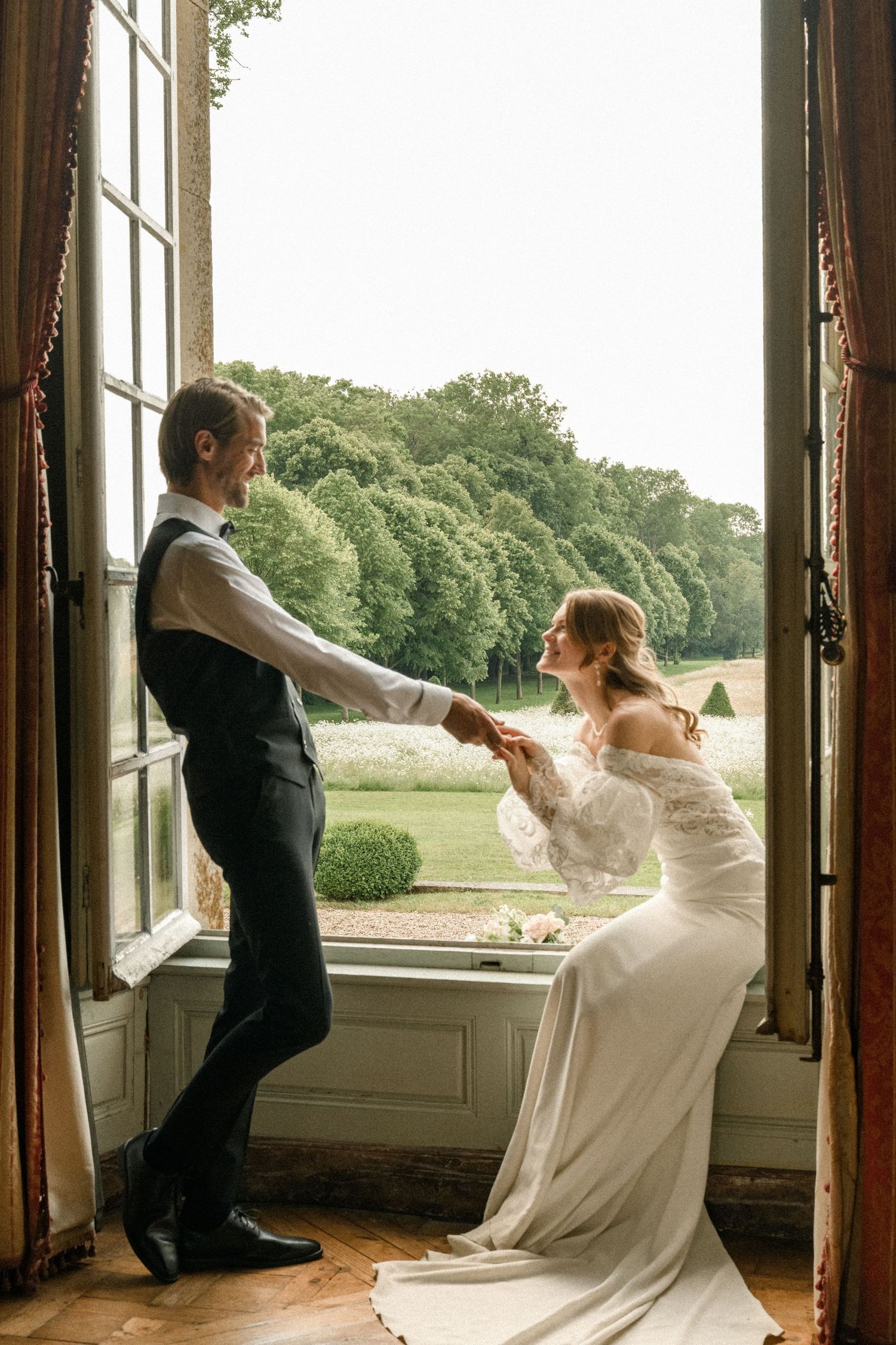 A couple in wedding attire dances playfully by a grand window in an elegant venue with garden views.