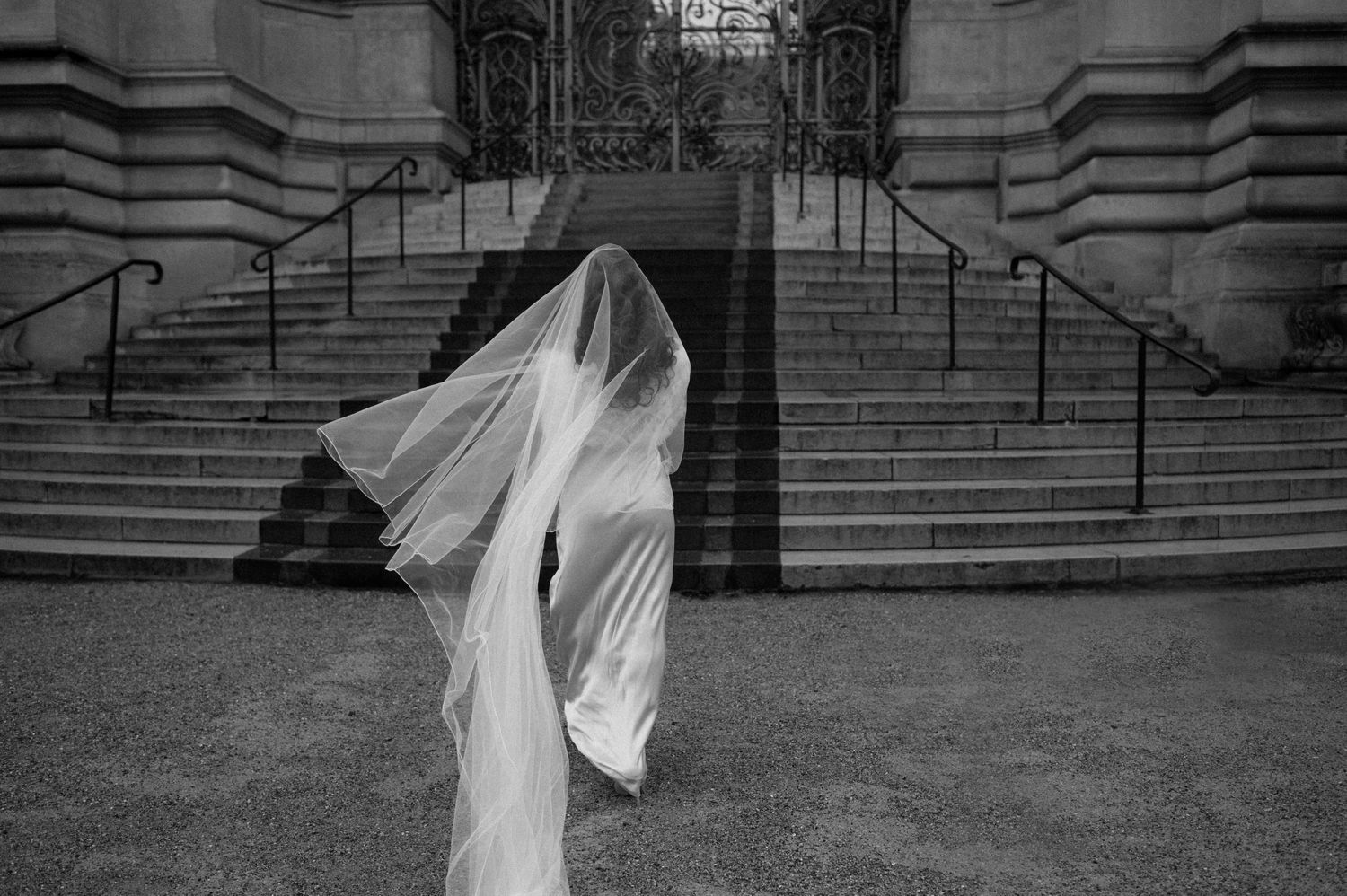 Artistic black and white photo of a bride's silhouette with flowing veil against stone steps.