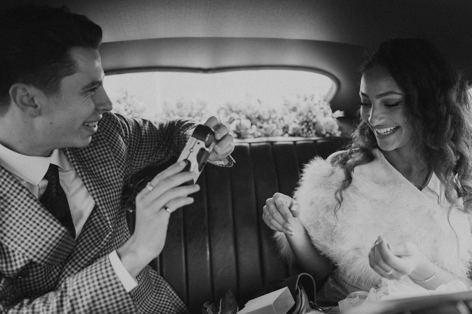 A black and white photo captures a playful moment between newlyweds in a vintage car.
