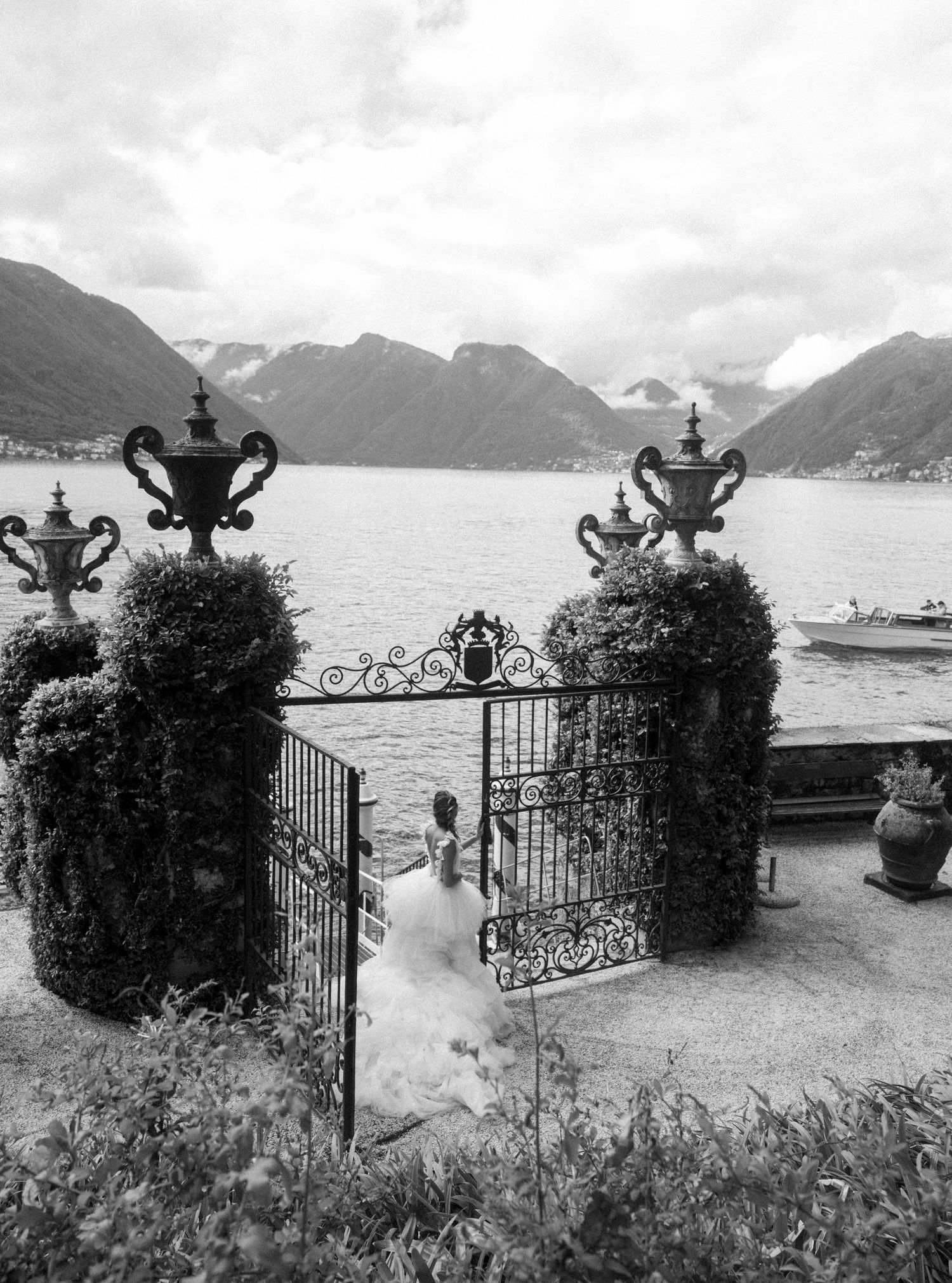 Black and white photo of ornate iron garden gates with ivy-covered pillars, urns, and a lake with mountains beyond.