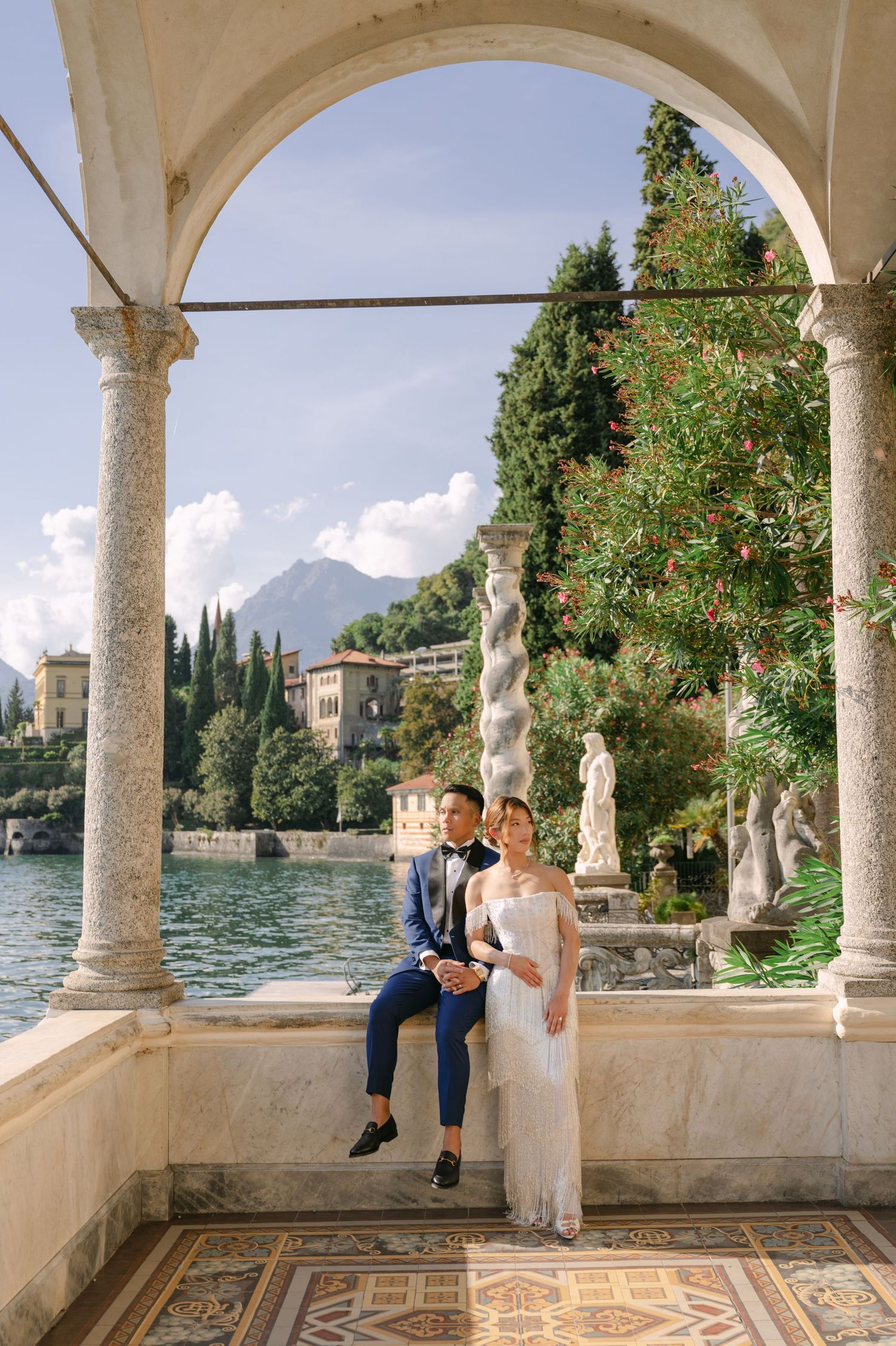 Bride and groom pose under elegant stone arches at Lake Como villa, mountains and lake visible behind them.