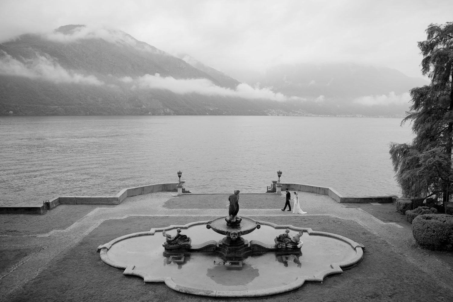 Black and white photo of a fountain terrace overlooking a misty lake with mountains in the background.