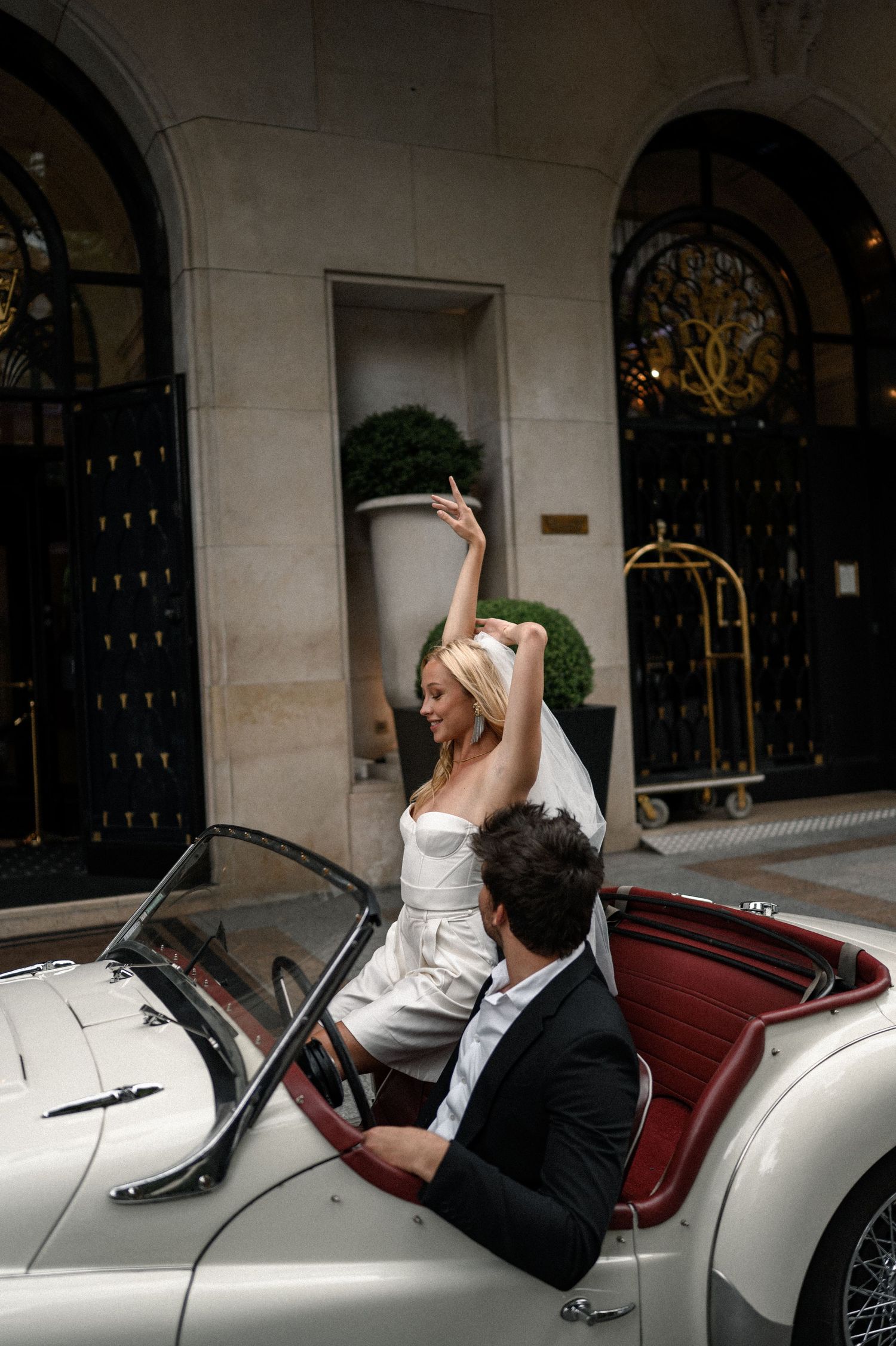 Bride in strapless white dress and veil celebrates in classic convertible with groom outside luxury Paris hotel.