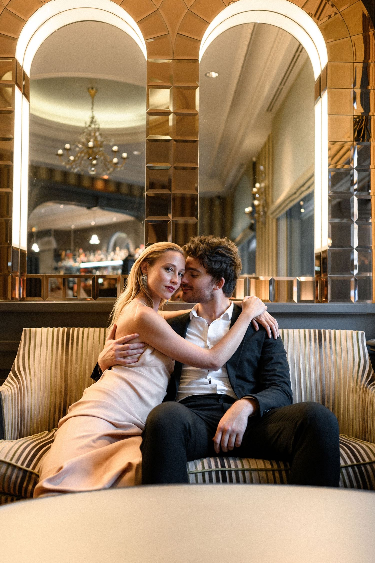 Romantic couple embracing on a striped sofa in a glamorous hotel lobby with golden decor.