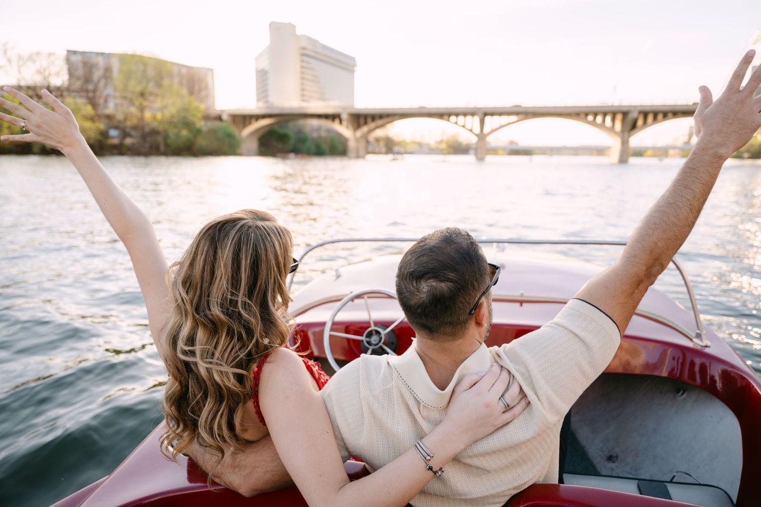 Couple with arms raised joyfully riding a classic red boat under a bridge.