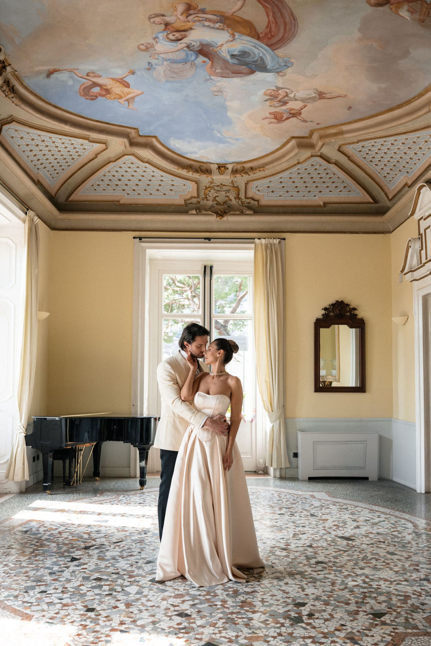 Couple shares romantic kiss in sunlit villa room with ornate baroque ceiling fresco and grand piano.