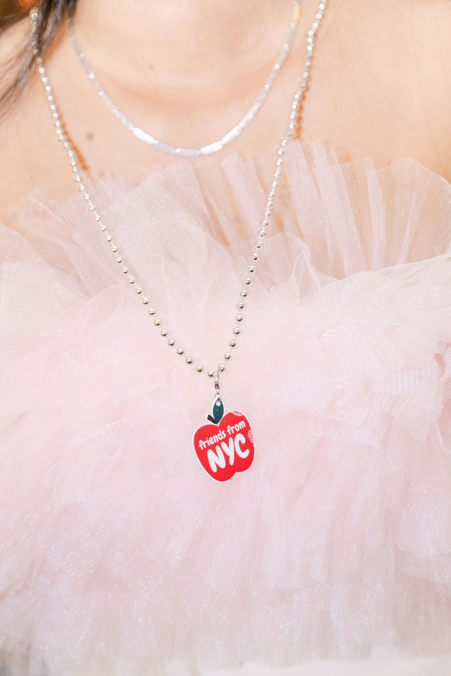 Close-up of a red NYC apple charm pendant on a silver ball chain necklace over a pink tulle dress.