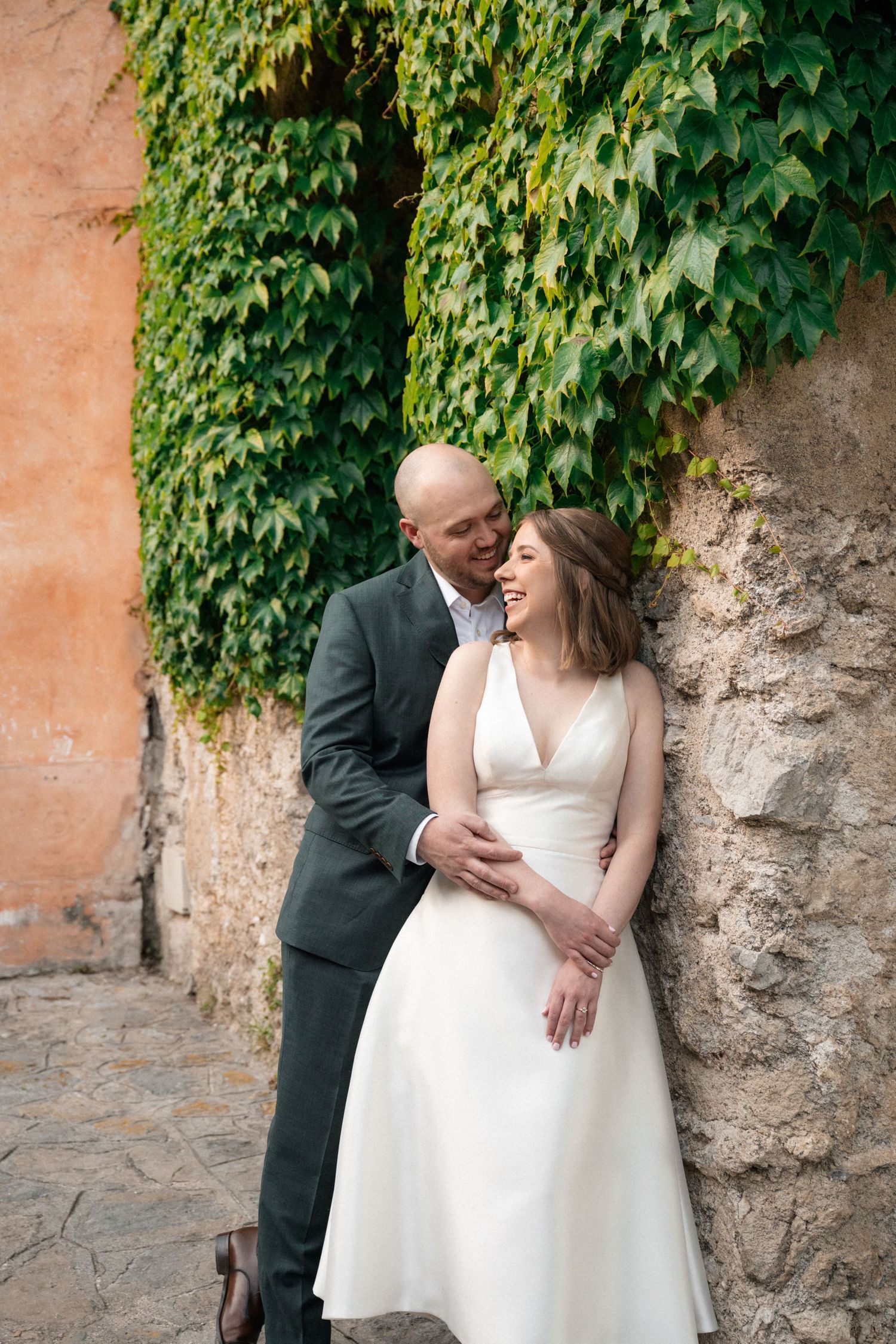 Groom embraces laughing bride against ivy-covered stone wall in romantic Italian wedding portrait.
