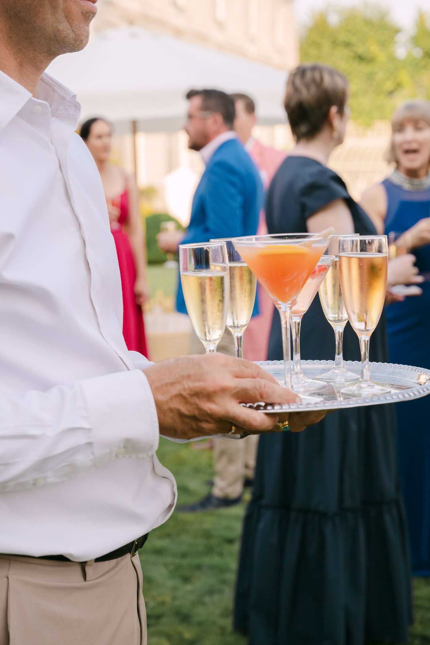 Waiter holding silver tray with champagne flutes and cocktails at elegant outdoor wedding reception.