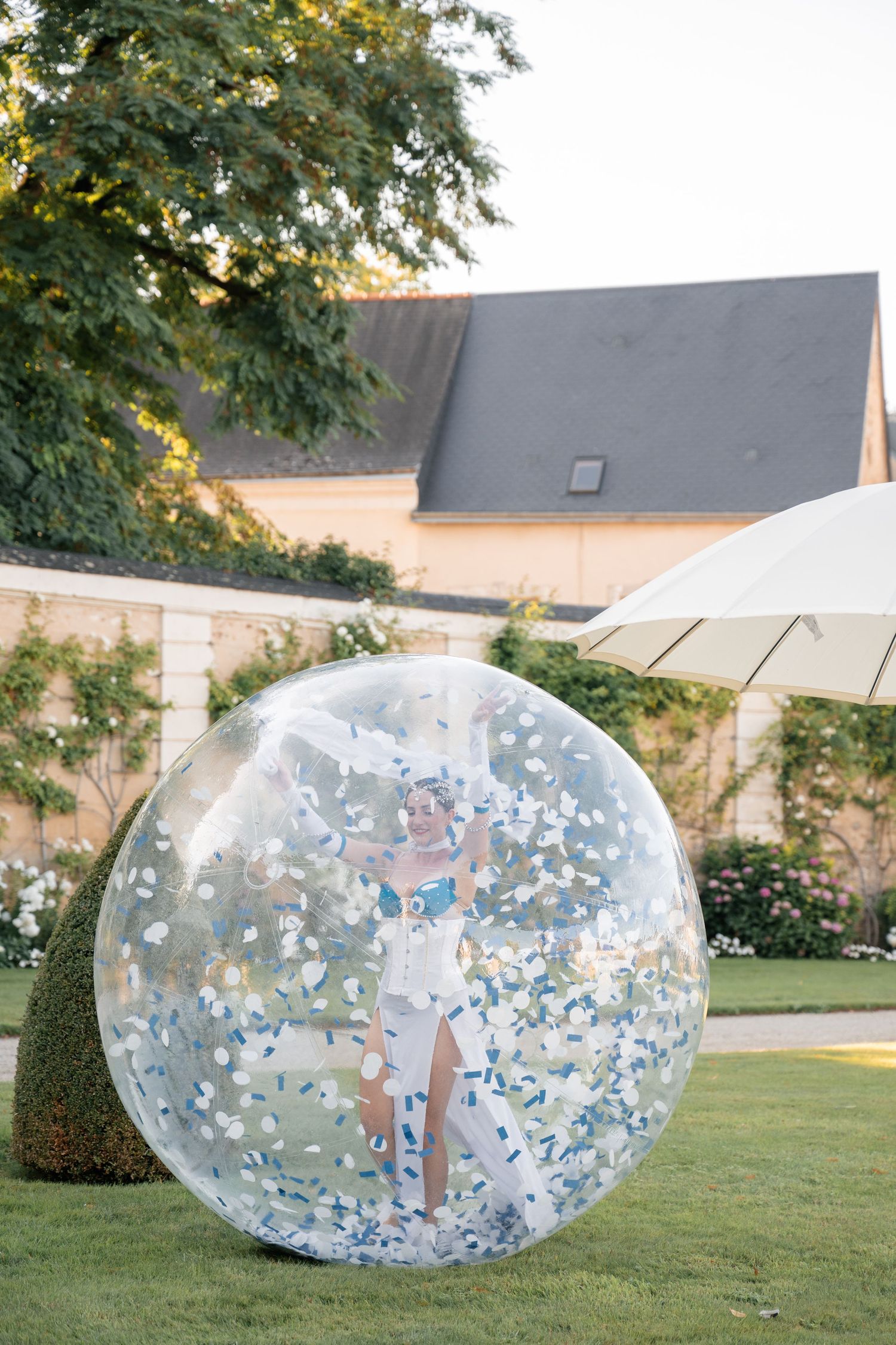 Performer inside giant transparent inflatable bubble filled with confetti at garden party.