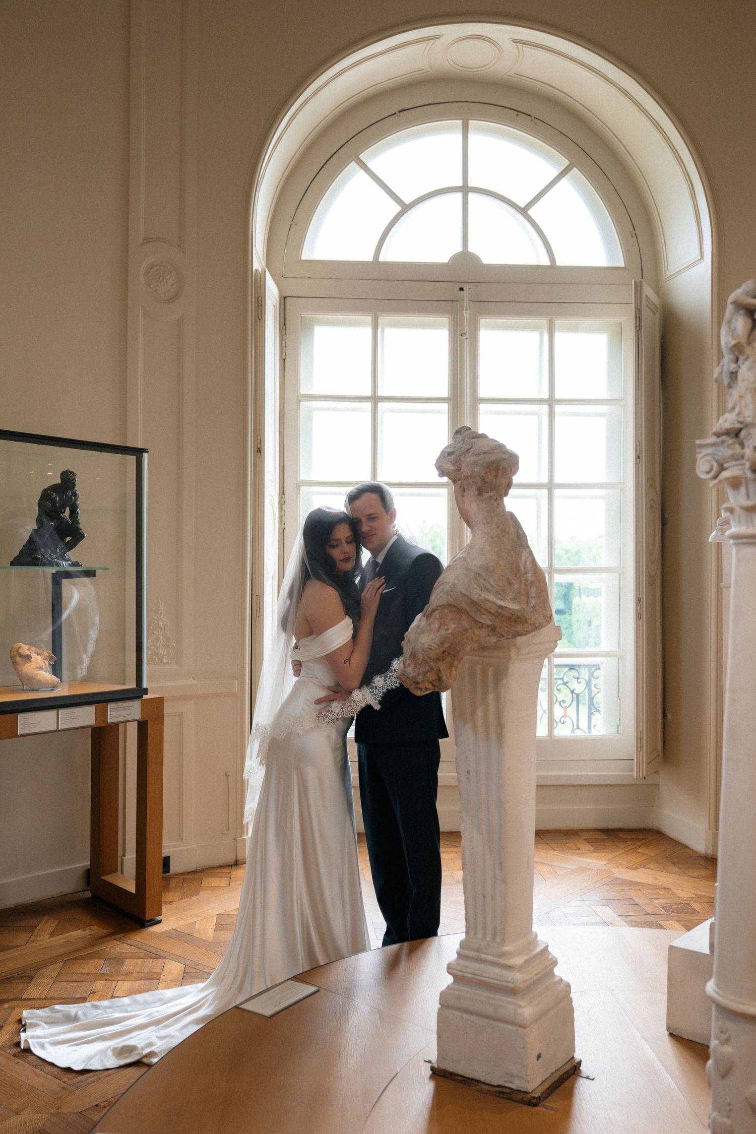 Bride and groom pose intimately beside an antique bust sculpture beneath an arched window in an elegant French museum.