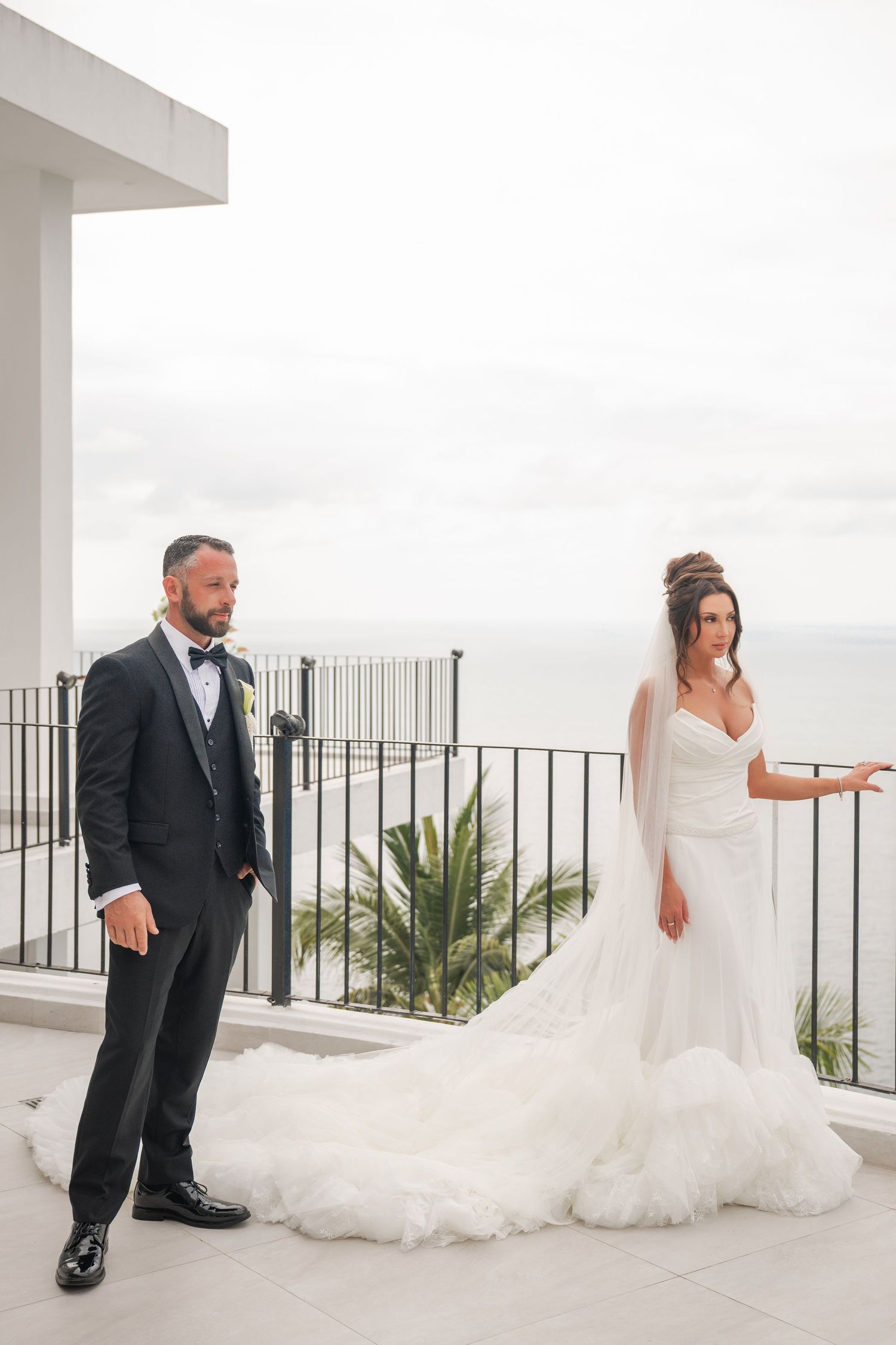 Bride in white ruffled gown with veil and groom in black tuxedo pose on seaside balcony.