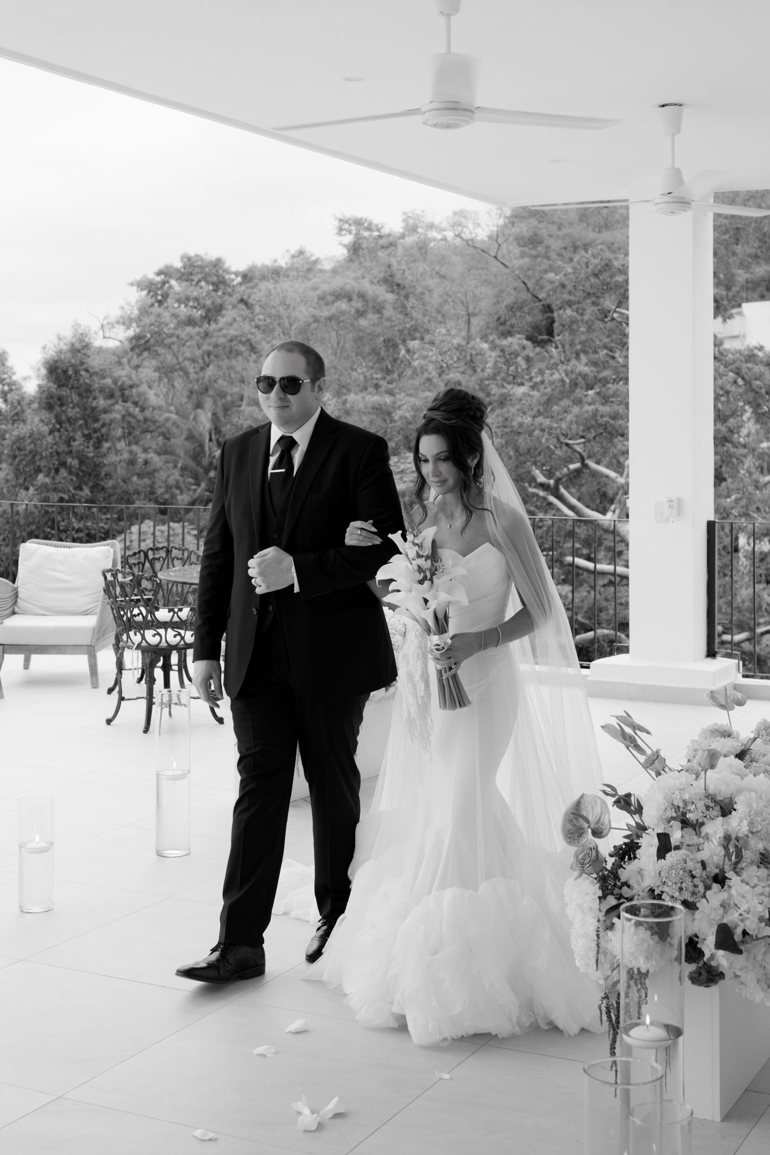 Bride and escort walking down aisle at outdoor wedding ceremony in black and white.