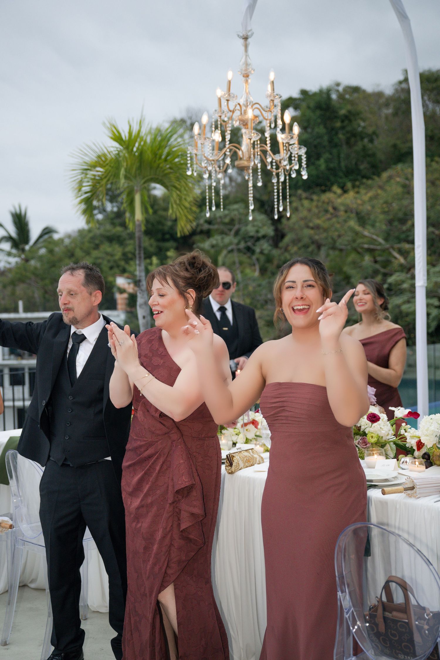 Bridesmaids in burgundy gowns laugh and clap at an outdoor wedding reception with chandelier and tropical backdrop.