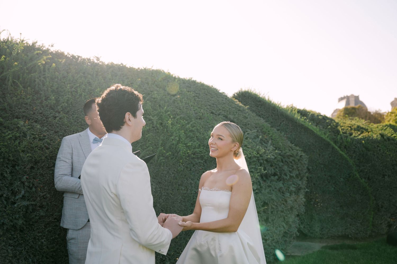 Bride and groom hold hands during outdoor wedding ceremony beside lush green hedge wall in golden sunlight.