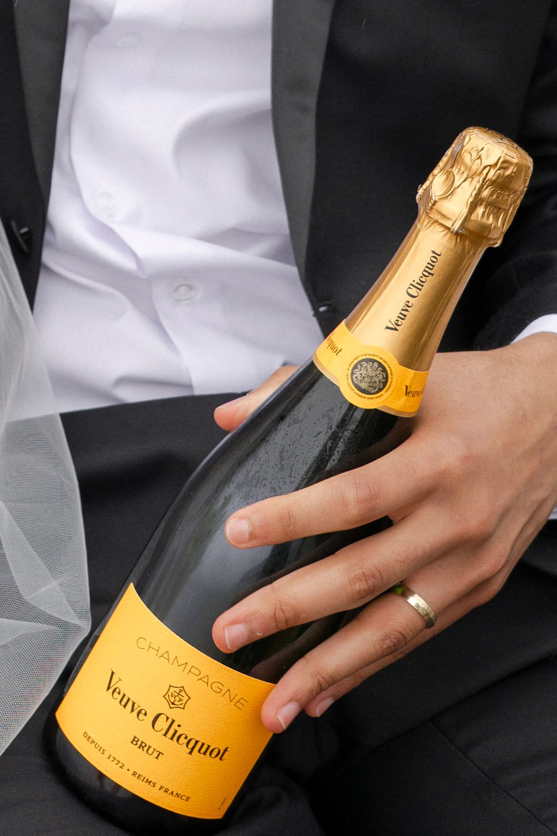 Groom in black tuxedo holds Veuve Clicquot champagne bottle with wedding ring visible on hand.