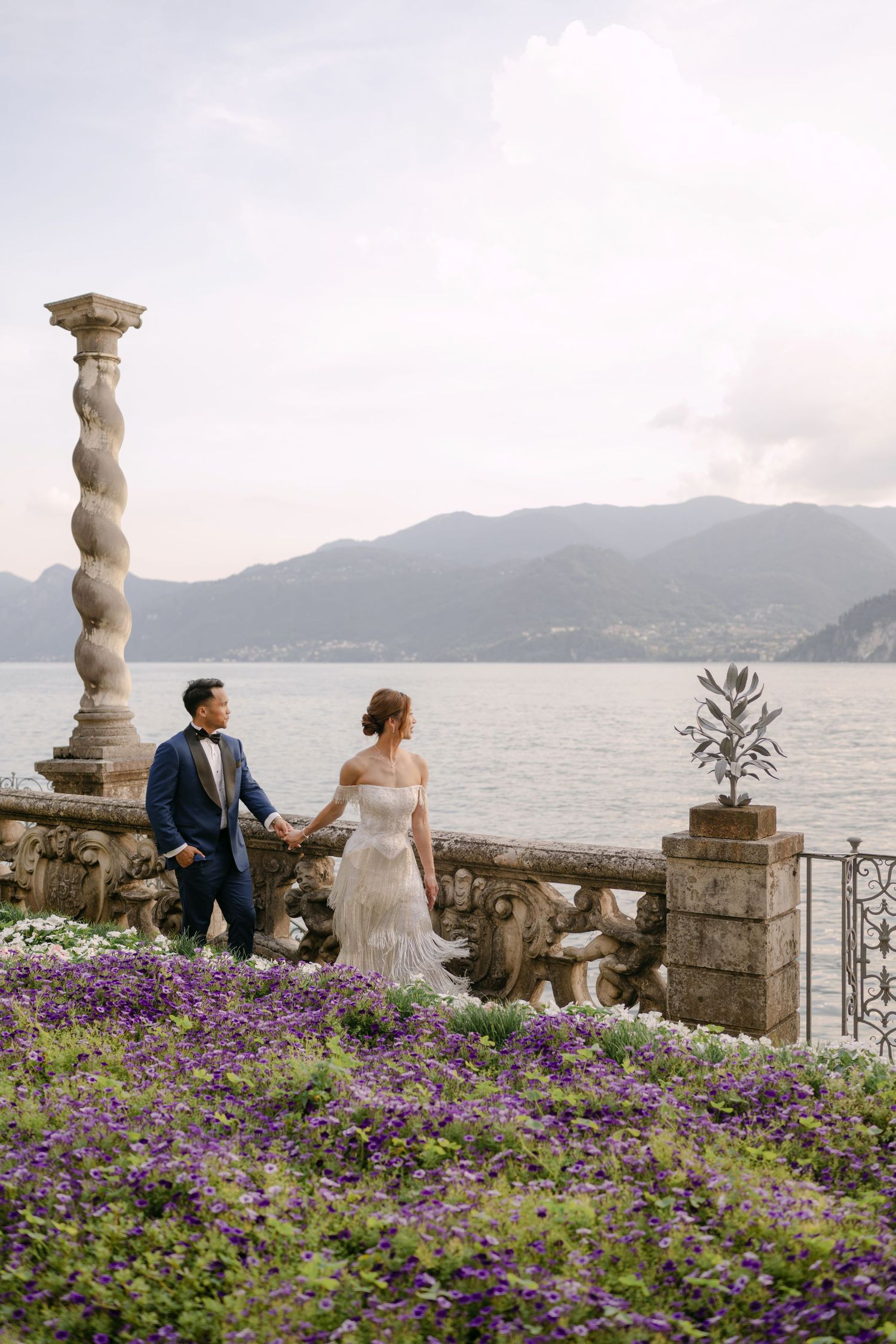 Bride and groom hold hands on a floral terrace overlooking Lake Como with mountain views in Italy.