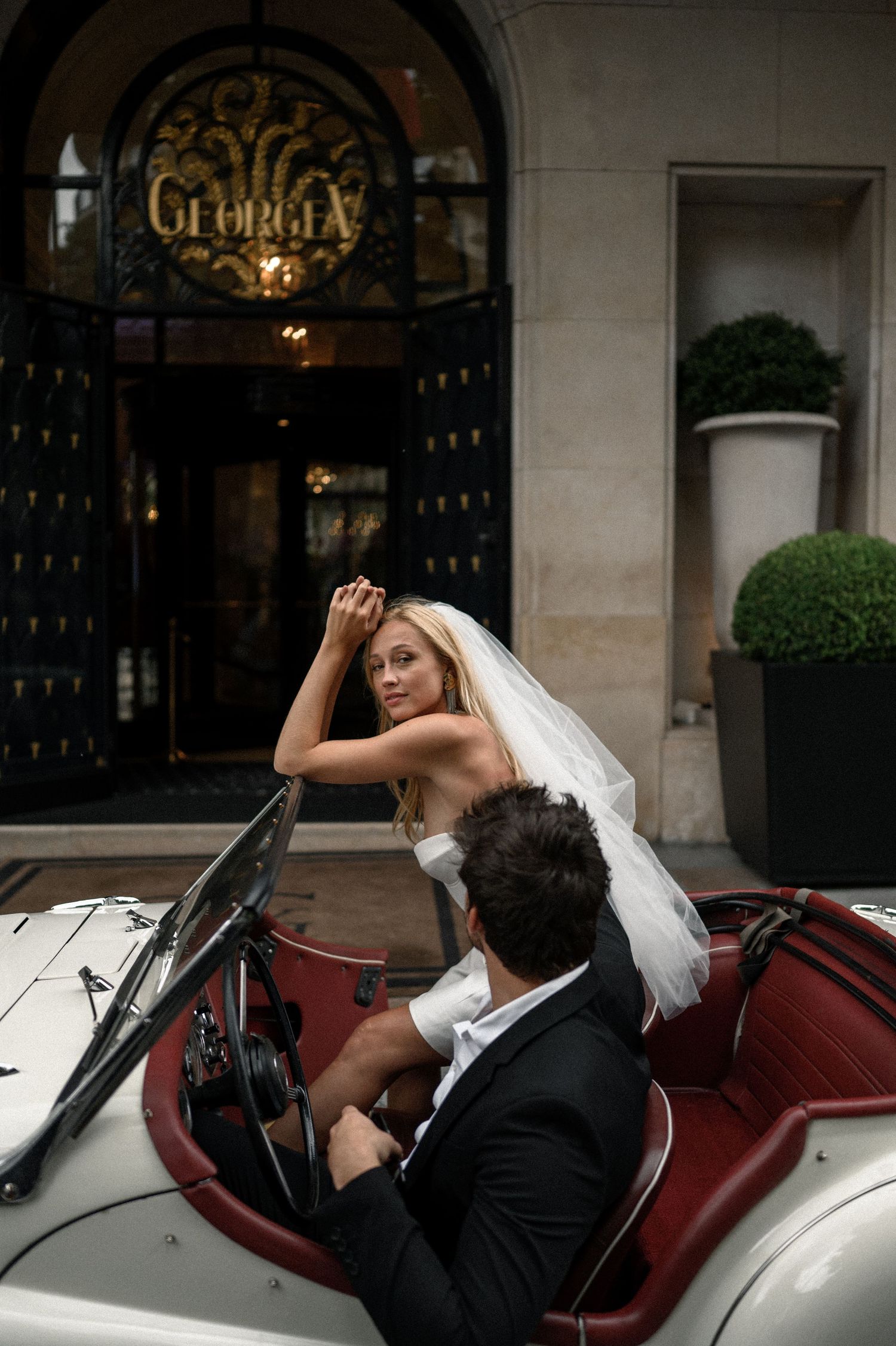 Bride in white strapless gown leans over vintage convertible windshield with groom outside Hotel Georgia Paris.