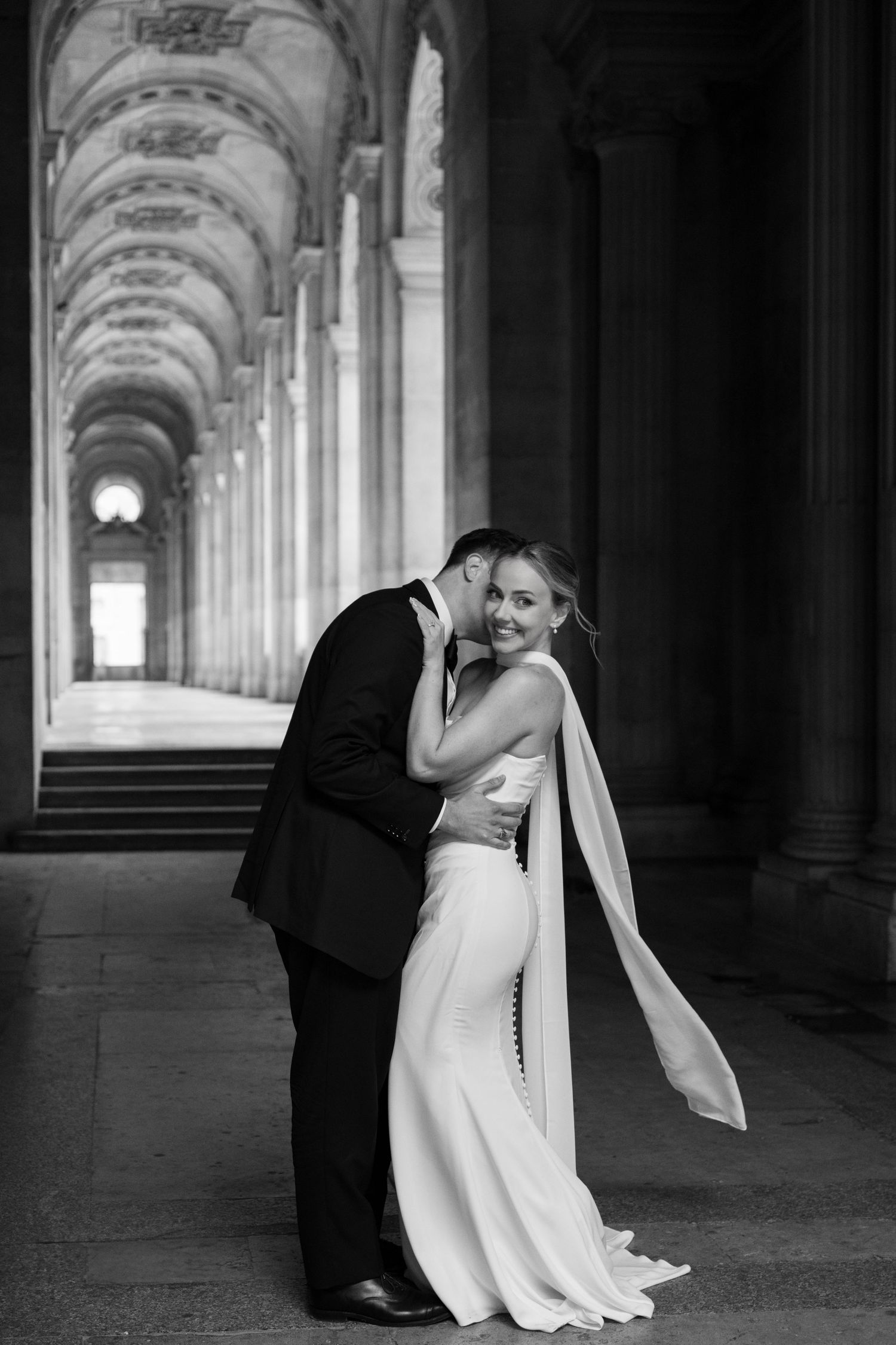 Black and white photo of groom kissing bride in a sleek white gown under ornate arched columns.