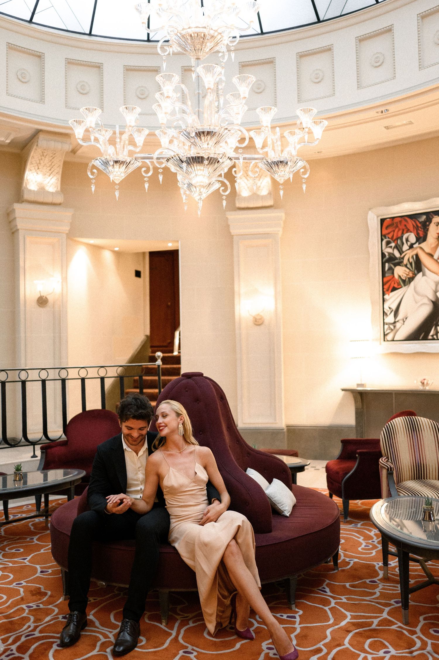 Couple relaxing on a velvet sofa beneath a grand chandelier in a classic hotel lobby.