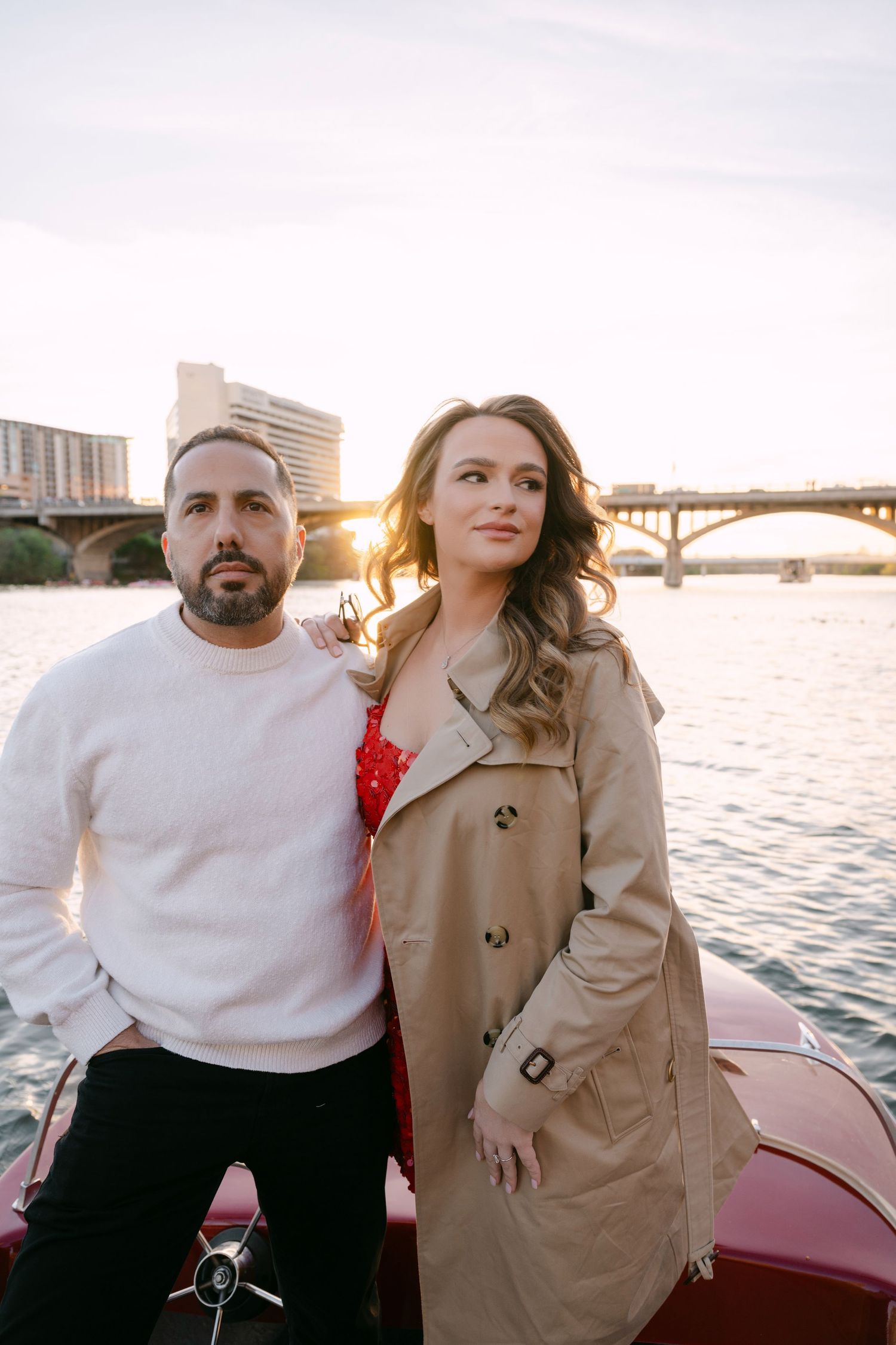 Couple posing on a red boat at sunset with a city bridge in the background.