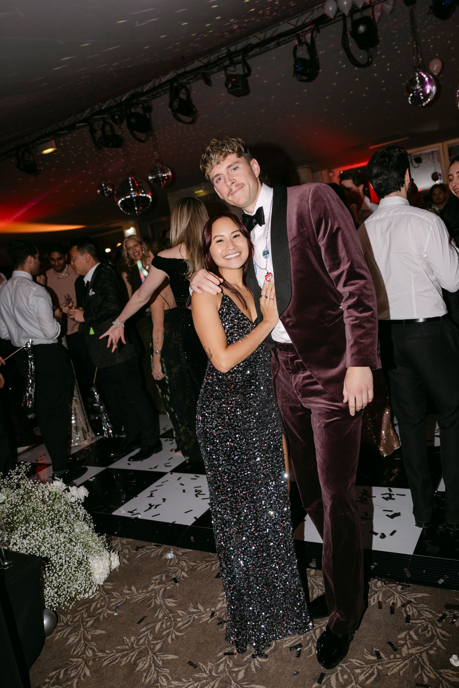 Couple posing together on dance floor at formal event, woman in black sequin gown and man in burgundy velvet suit.