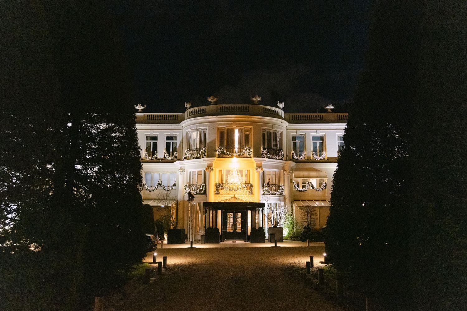 An illuminated grand mansion venue glows against the night sky, surrounded by manicured trees and a lit pathway.