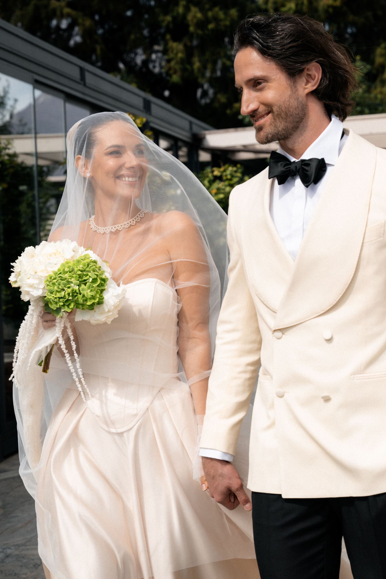 Smiling bride in veil holds green and white bouquet while walking hand in hand with groom.