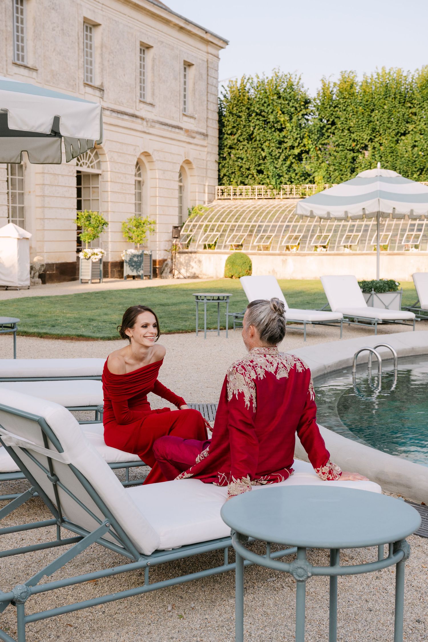 Two women in red dresses sitting on lounge chairs by pool at elegant French chateau estate.