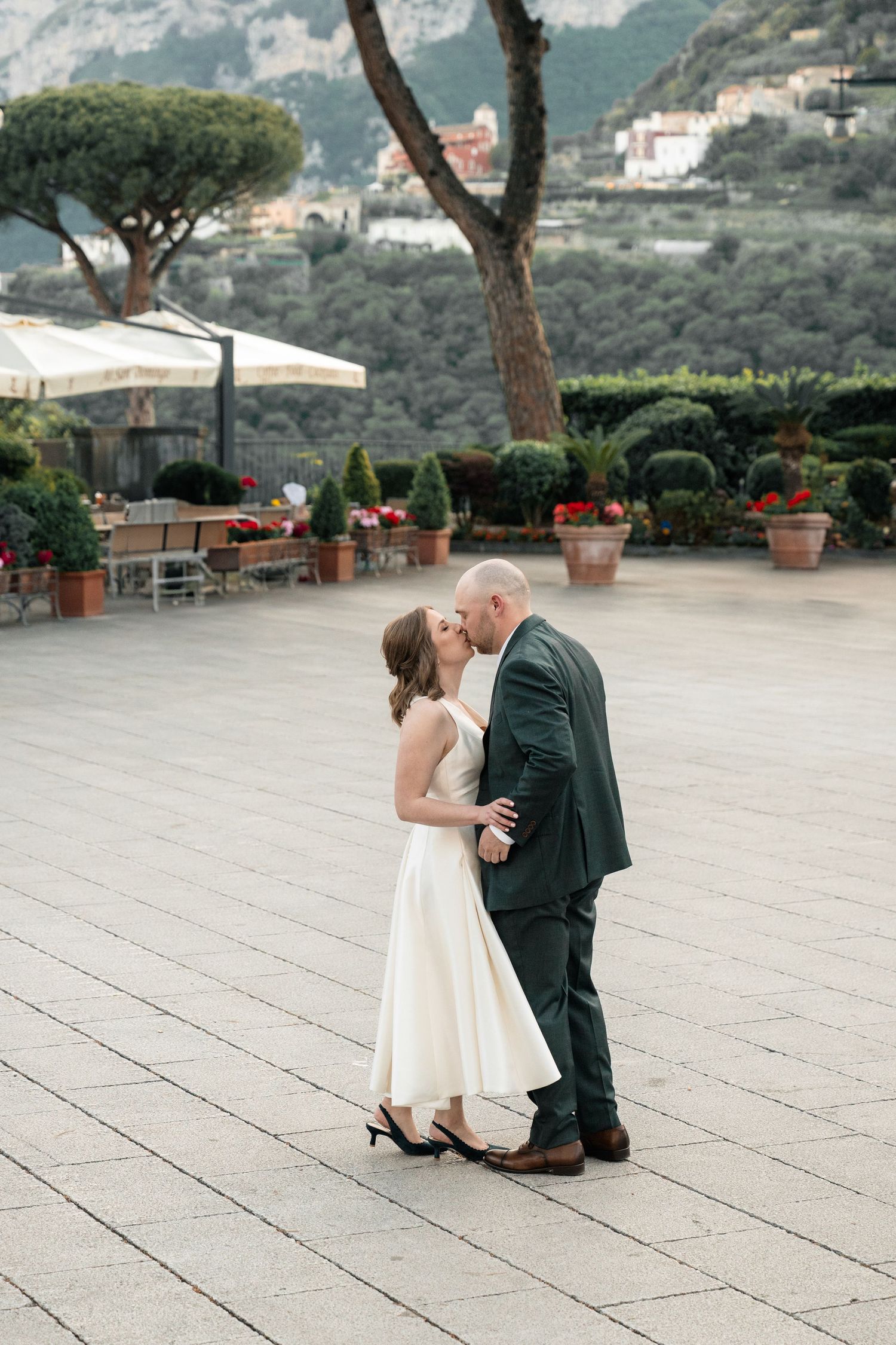 Bride and groom share a kiss on a stone piazza with Italian hillside garden landscape behind them.