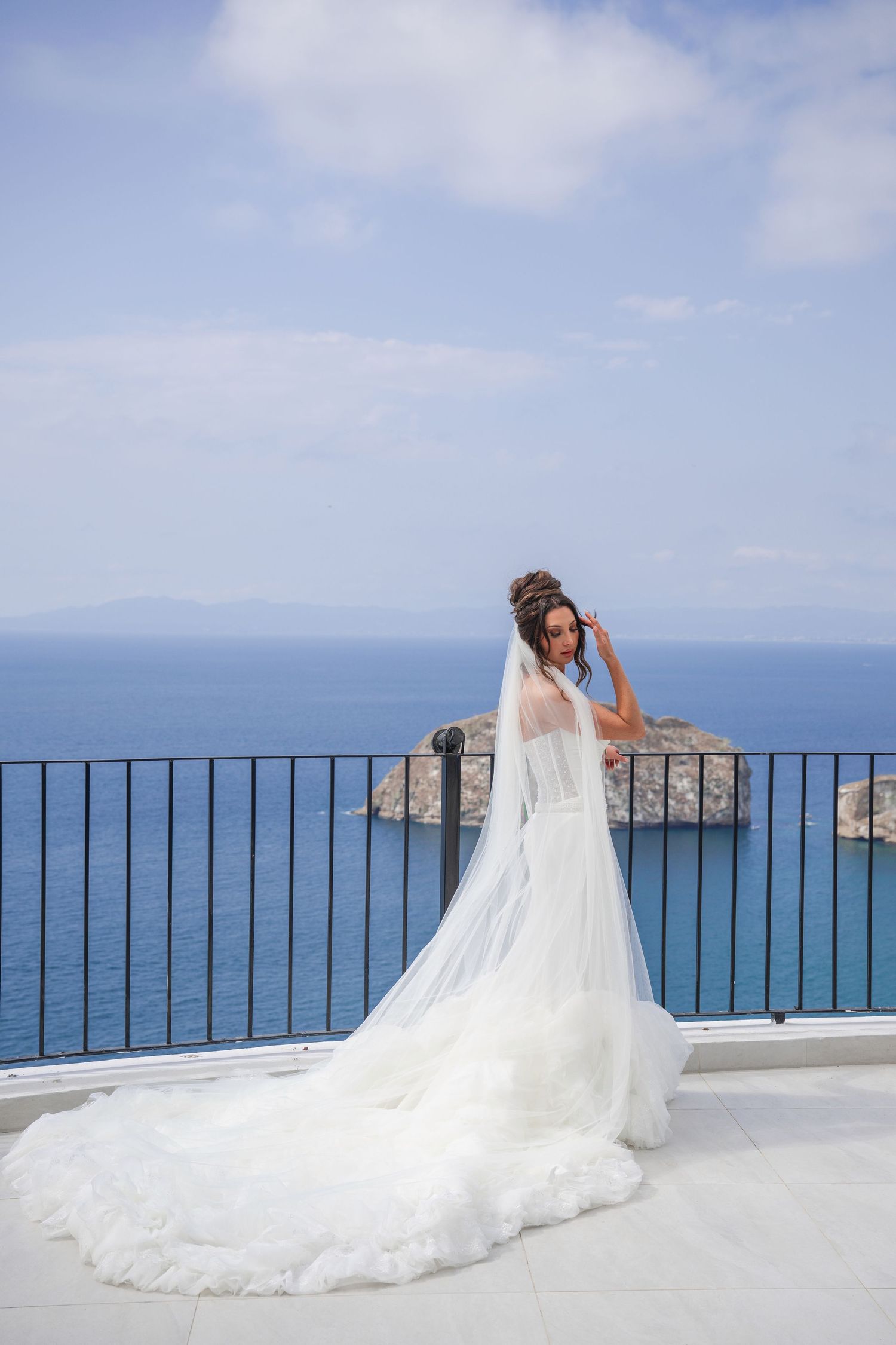 Bride in stunning white wedding gown with long veil stands on terrace overlooking blue ocean vista.