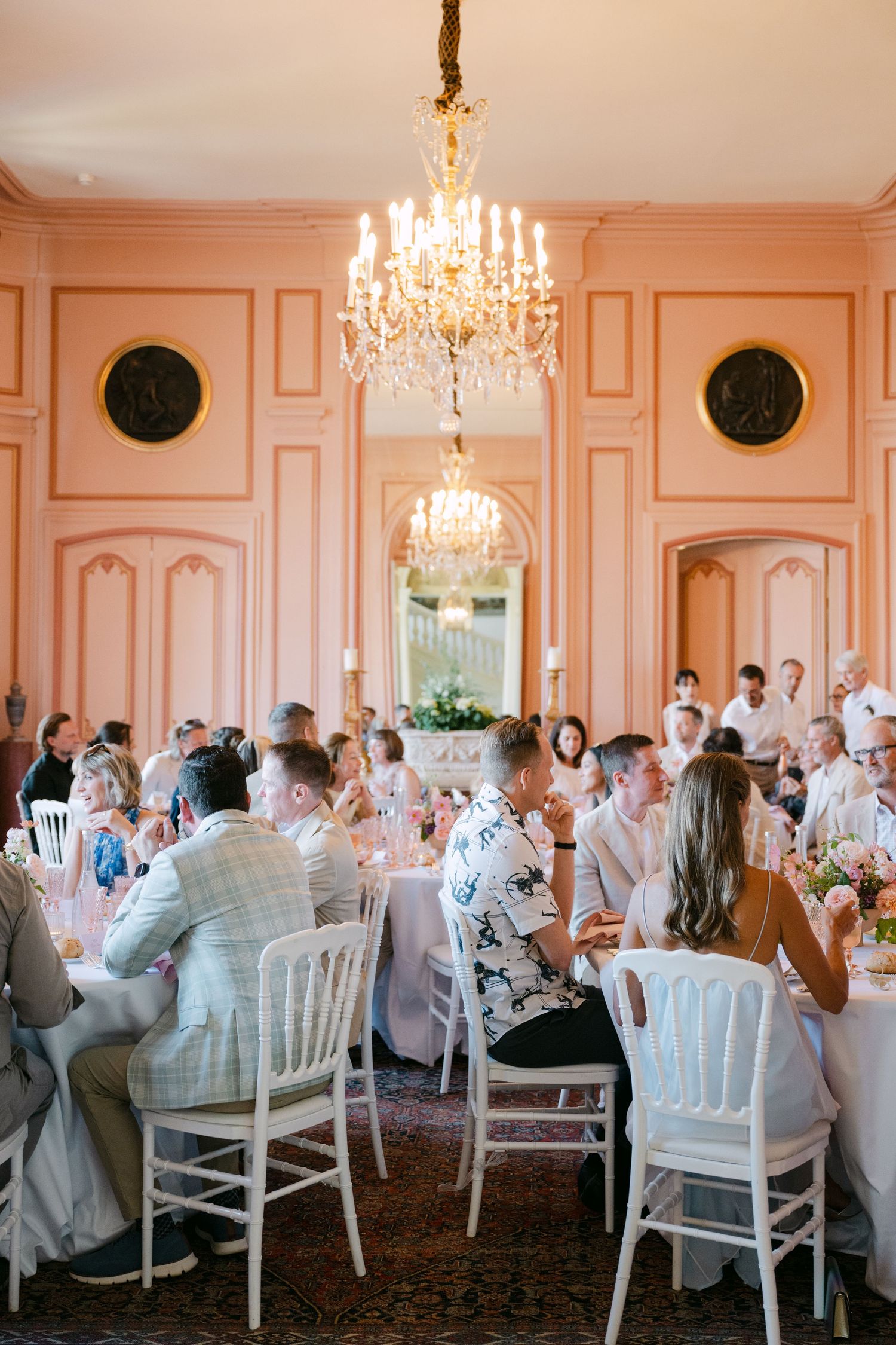 Wedding guests seated at round tables beneath a crystal chandelier in a grand pink French chateau reception hall.