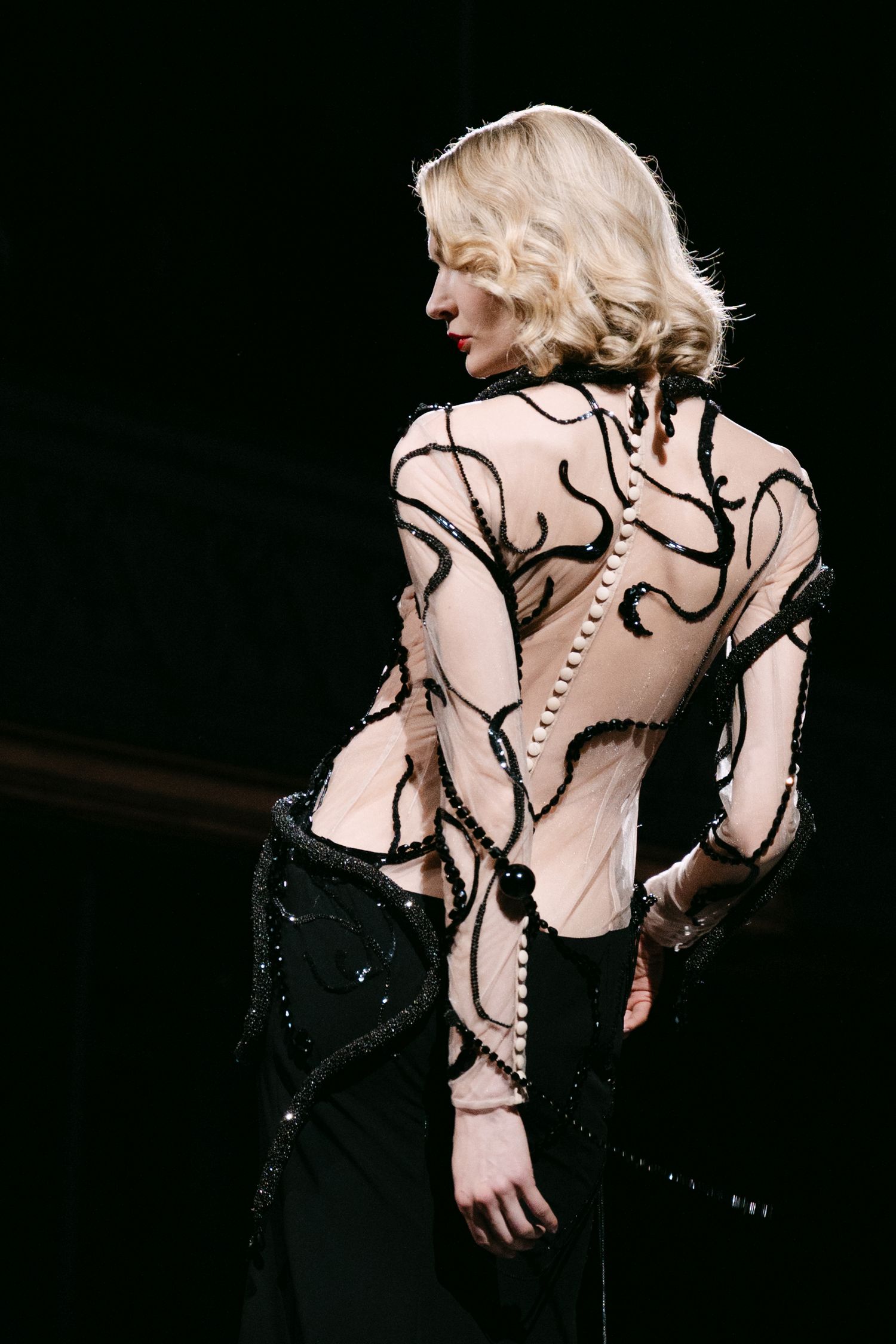 Model showcases sheer bodysuit with black swirl embroidery and sequined skirt on runway.