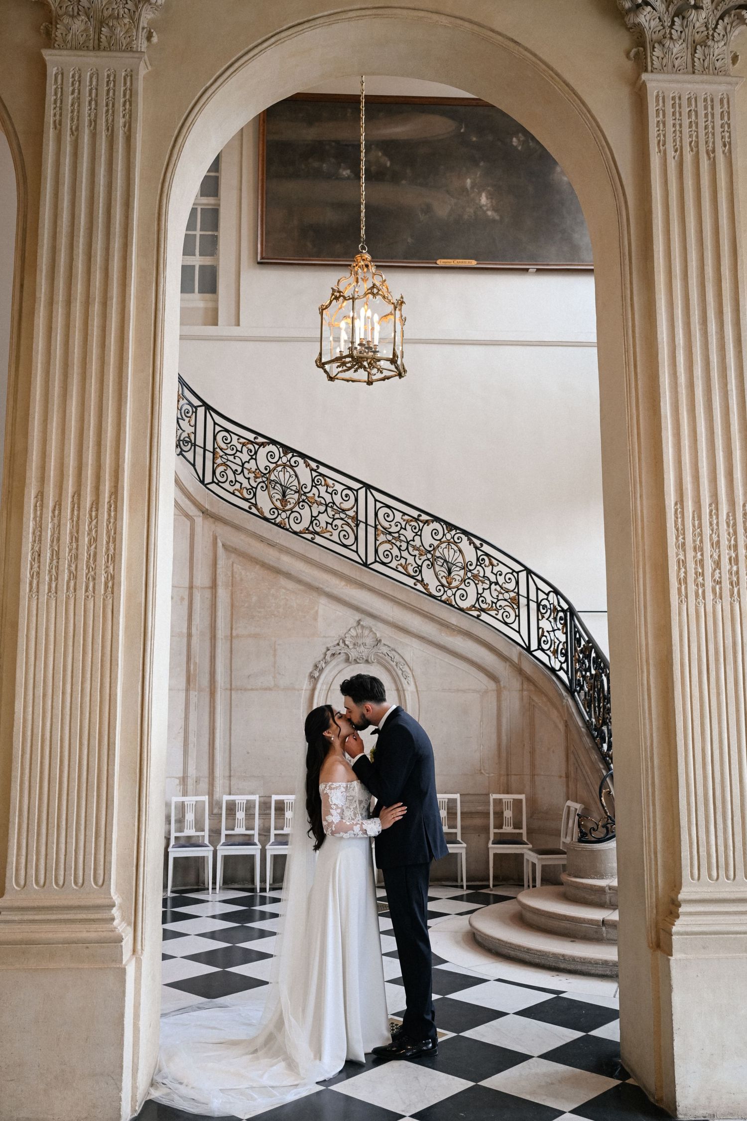 Bride and groom share tender embrace framed by ornate arch with spiral staircase in grand French chateau.