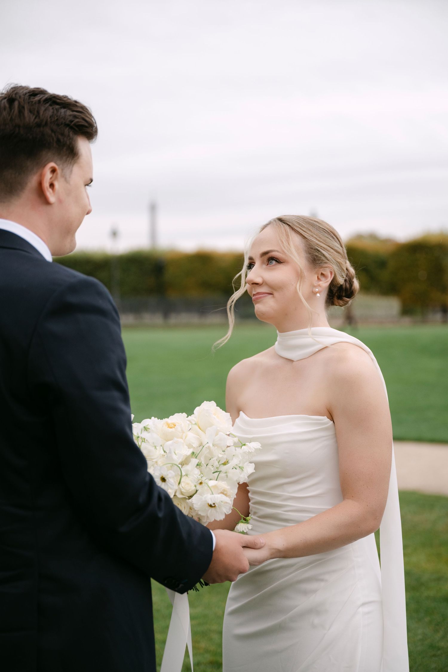 A couple in wedding attire exchange flowers during an outdoor ceremony with green fields in the background.