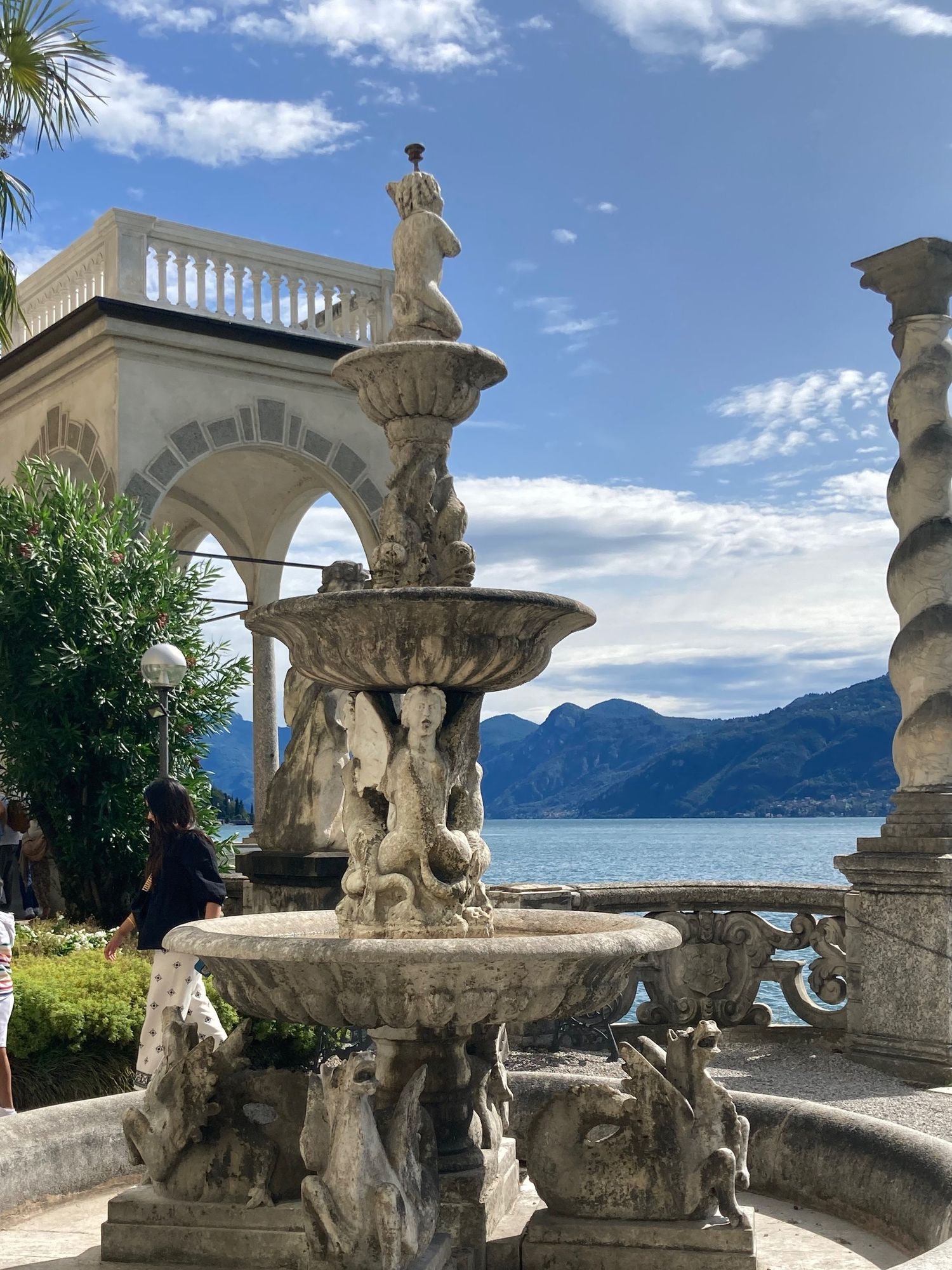 An elaborate tiered fountain with stone sculptures against a backdrop of mountains and lake.