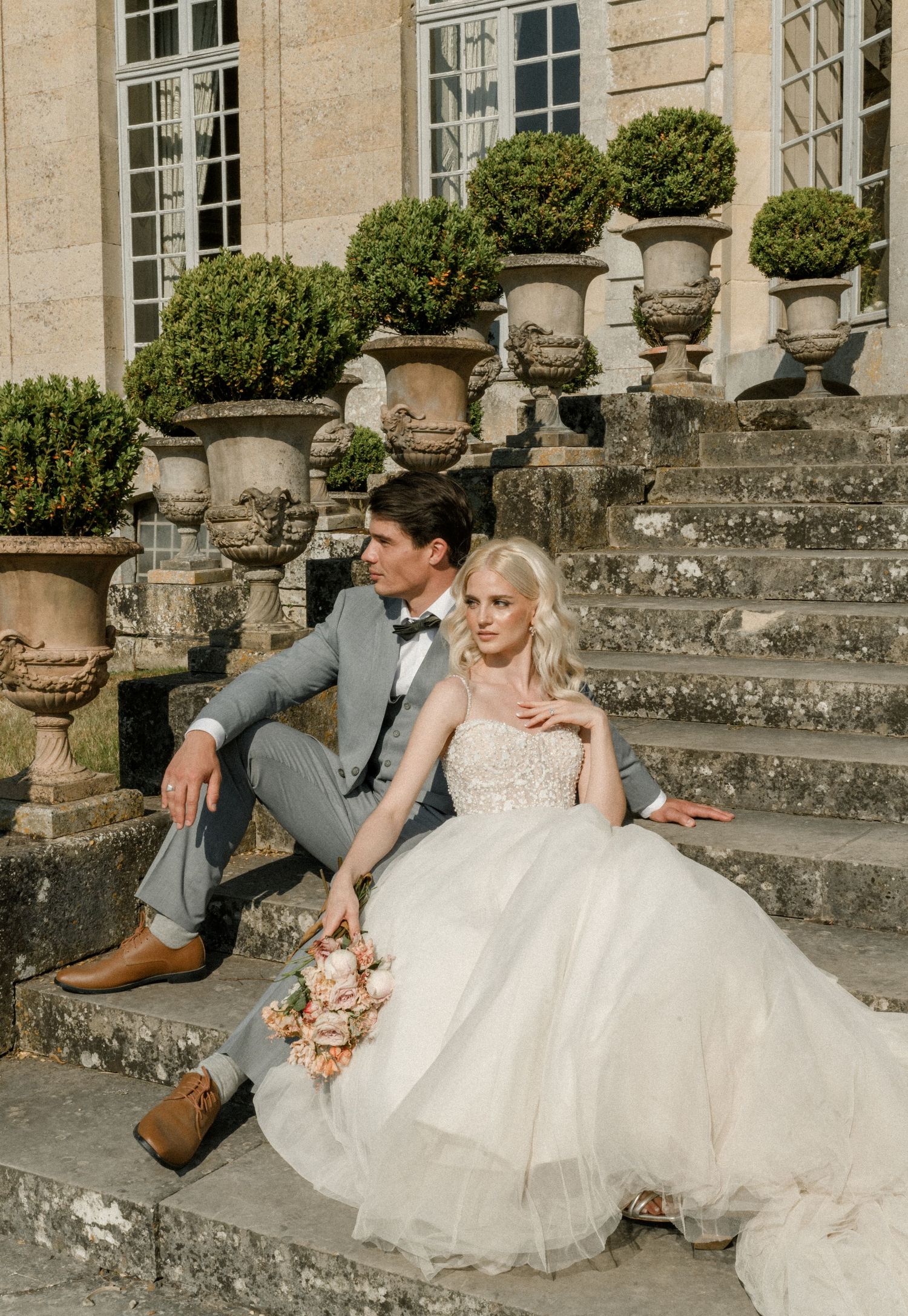 A wedding couple sits on ornate stone steps adorned with classical garden urns and topiaries.