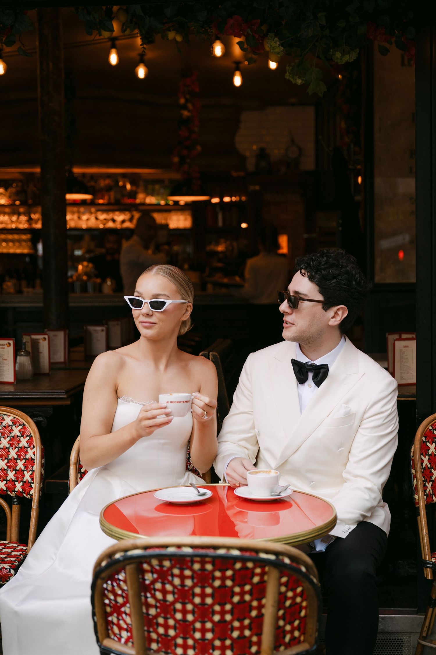 Stylish bride and groom wearing sunglasses enjoy coffee at a classic Parisian cafe with red bistro furniture.