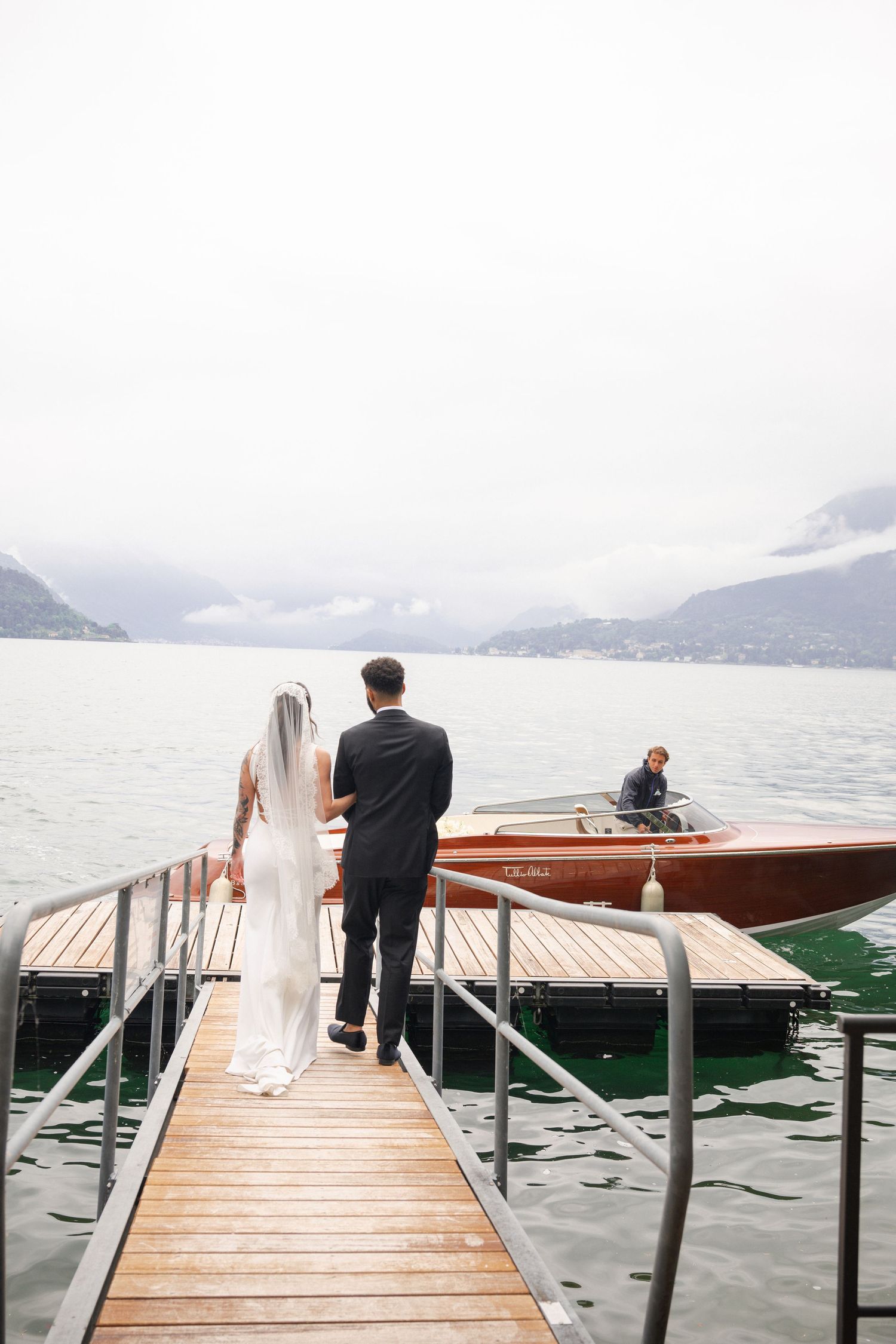 Bride and groom walk hand-in-hand on wooden dock toward classic wooden boat on misty alpine lake.