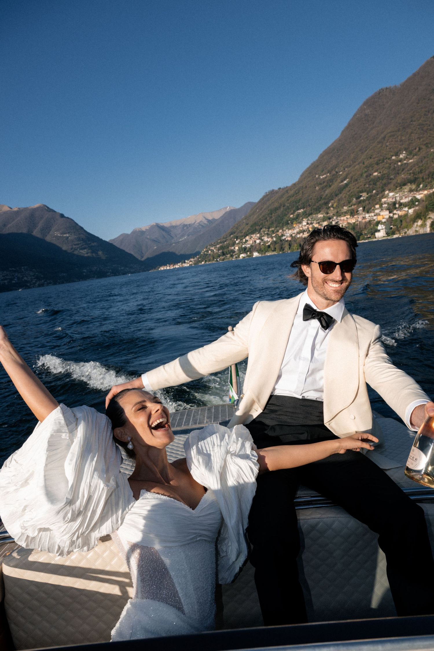 Joyful bride and groom celebrate on a luxury boat on Lake Como, Italy, with stunning mountain scenery under blue skies.