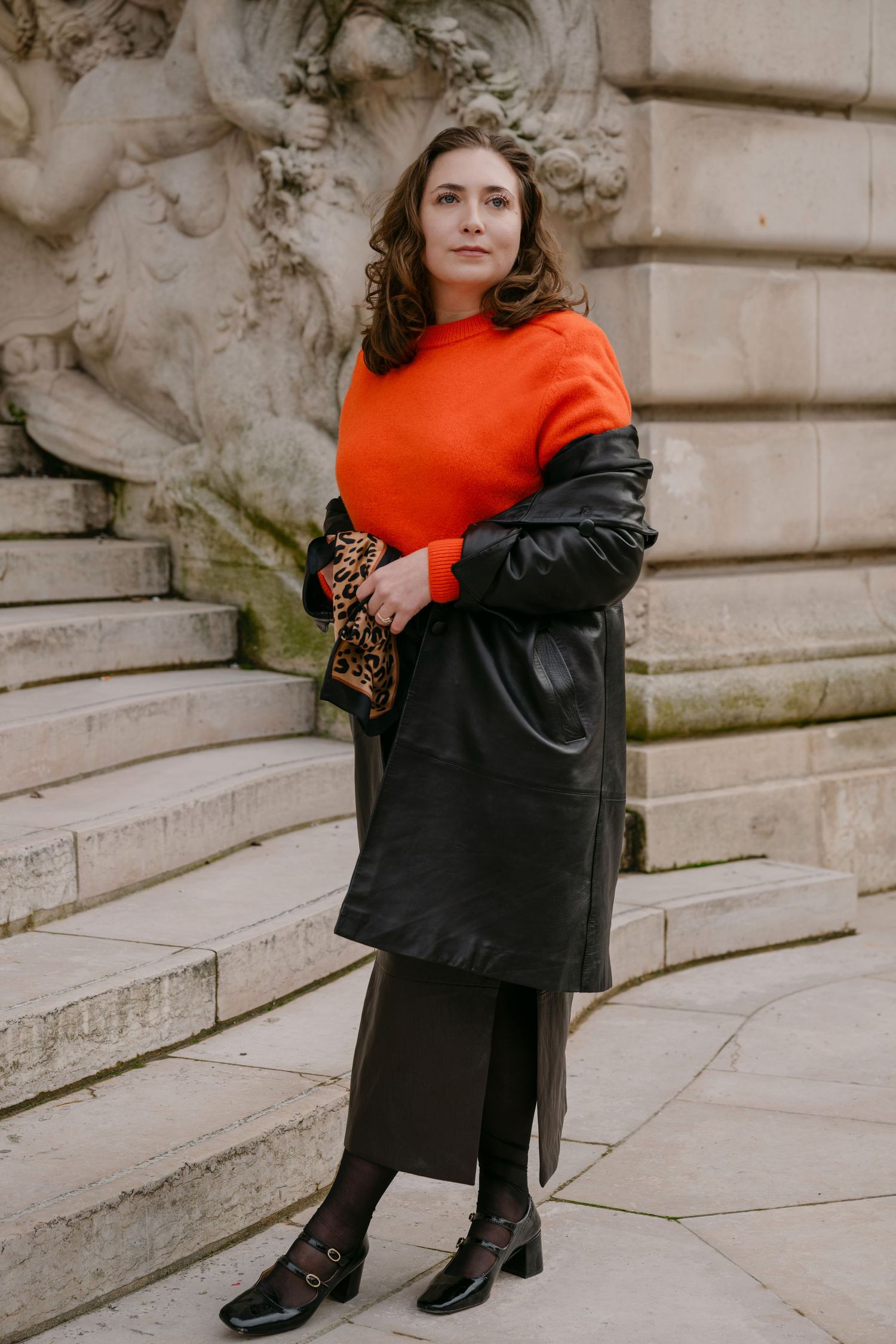 Woman in orange sweater, black leather coat, dark skirt, and heels, holding leopard clutch on stone steps.