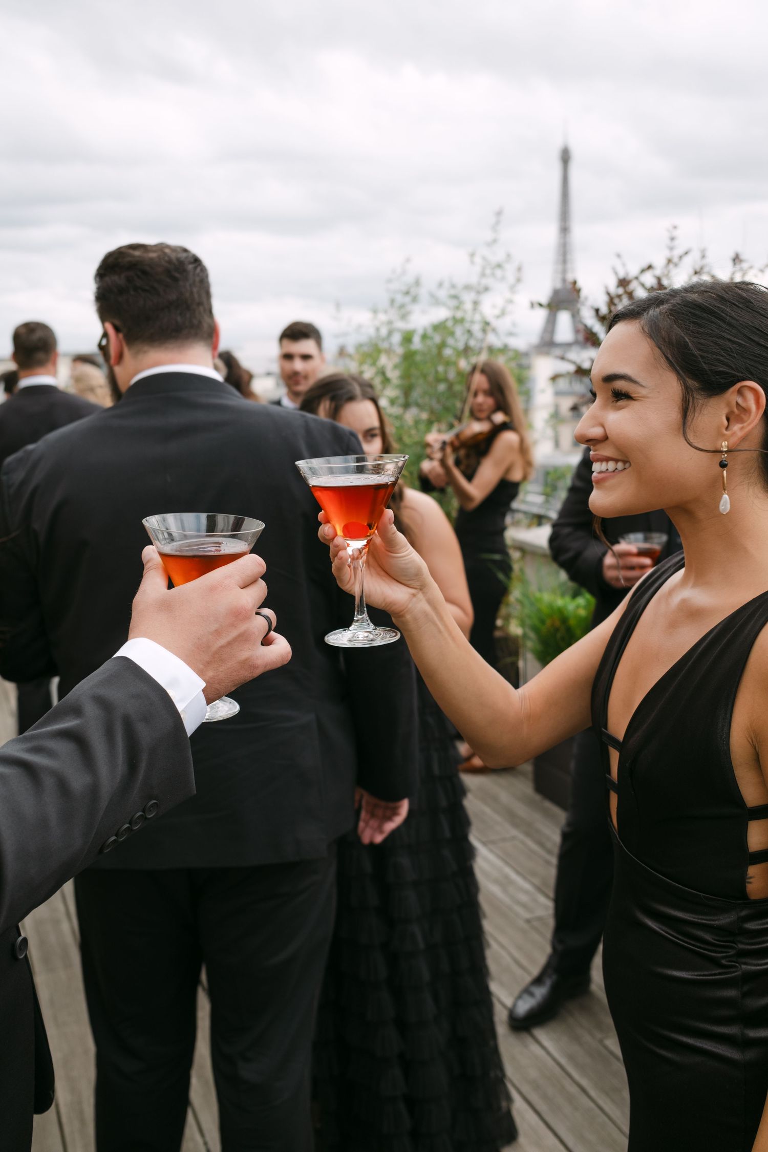 Guests in black formal attire toasting with cocktails at a rooftop party with the Eiffel Tower in background.