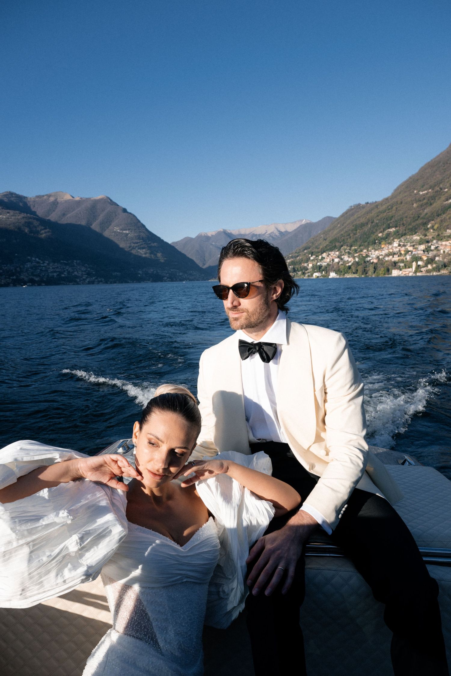Elegant bride and groom in wedding attire on a luxury boat on Lake Como with mountain backdrop.