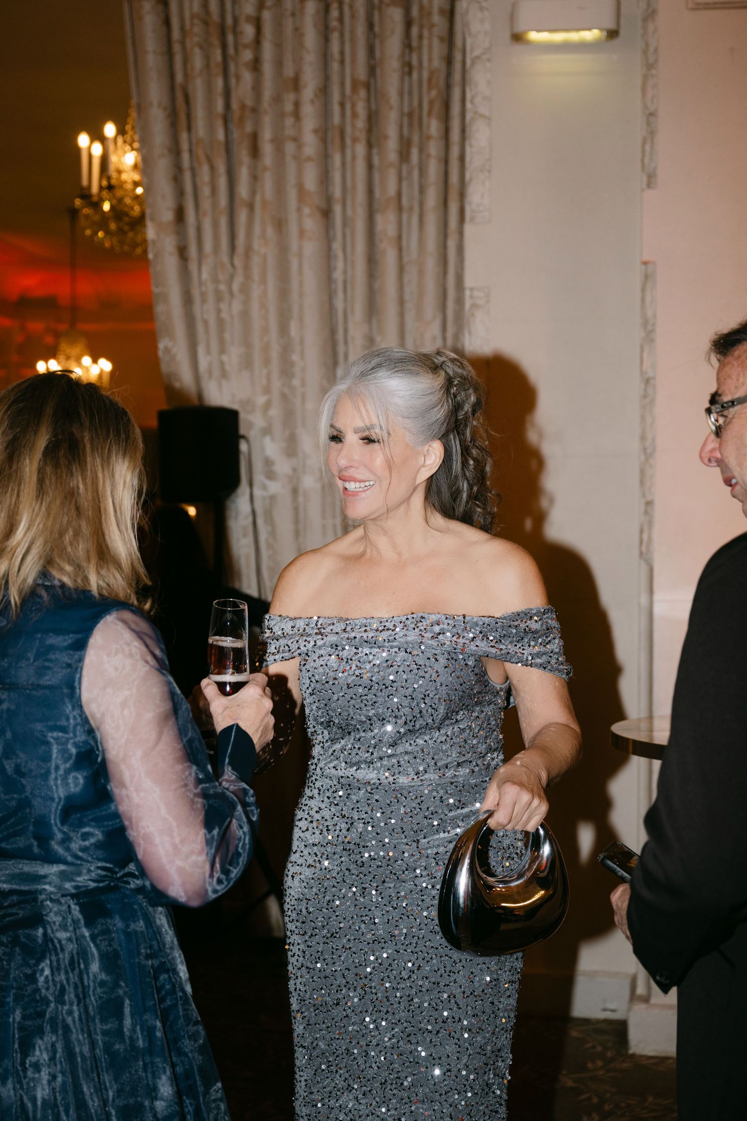 Elegant older woman in silver off-shoulder sequin gown holding black clutch at formal evening event.