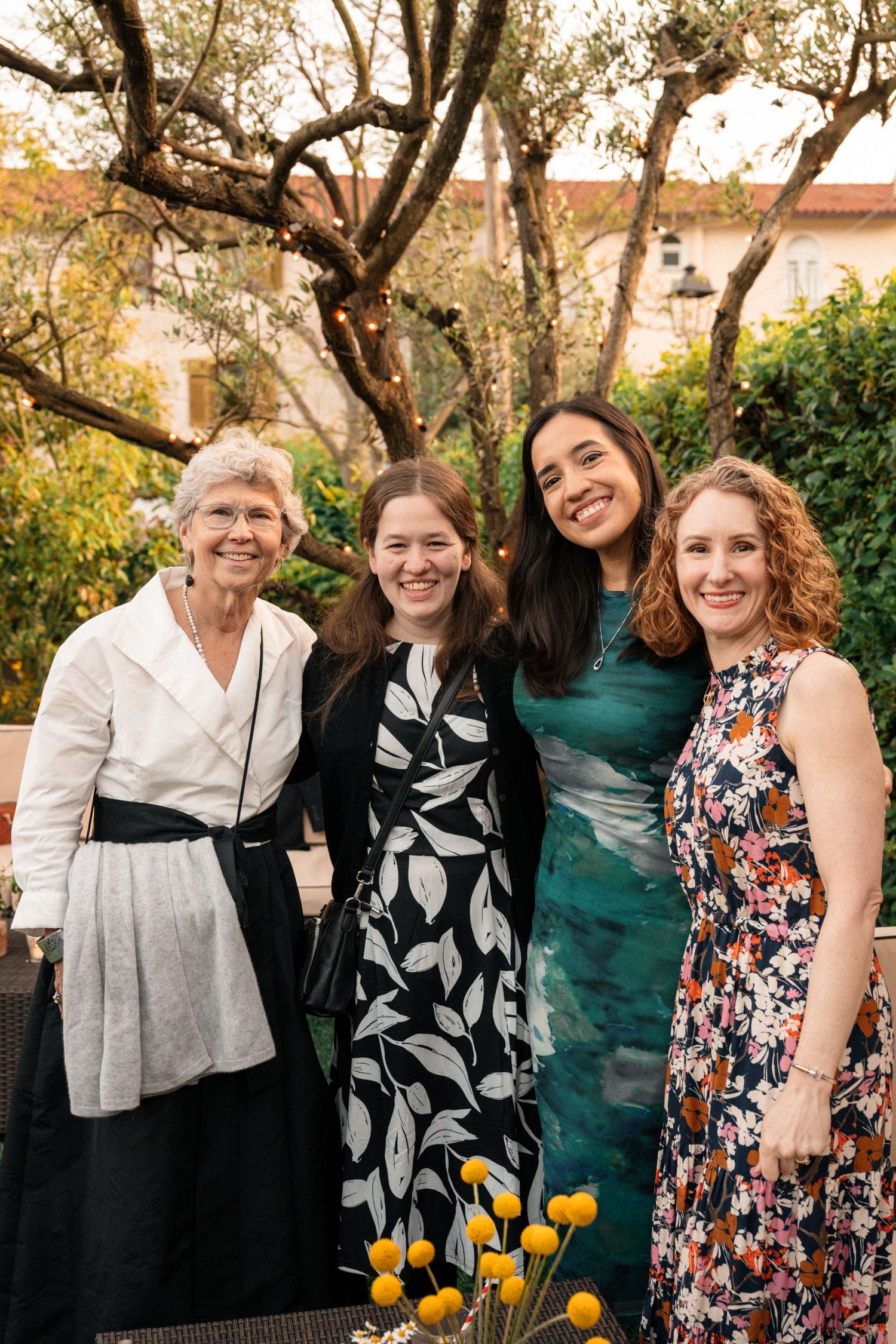 Four women smiling together at outdoor garden wedding reception beneath olive trees with yellow flowers.