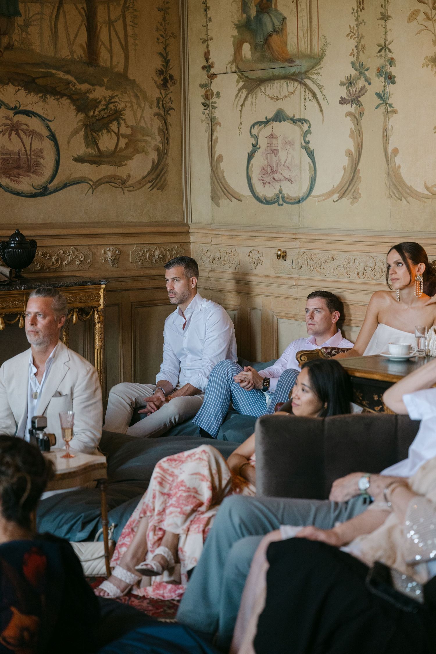 Wedding guests attentively seated in a luxurious French chateau salon with ornate painted wall murals.