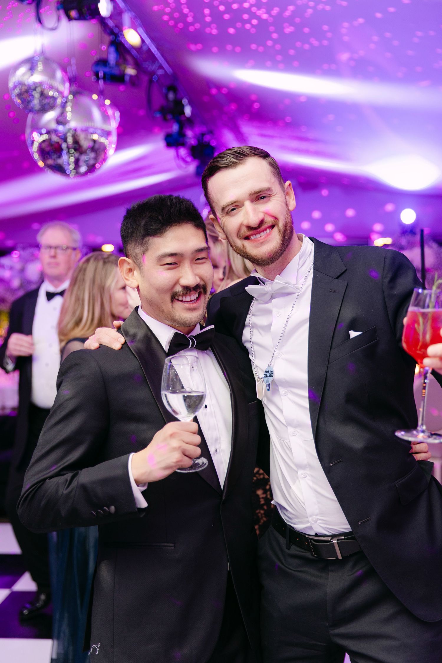 Two men in tuxedos smile and hold champagne glasses at an elegant wedding reception with purple lighting.