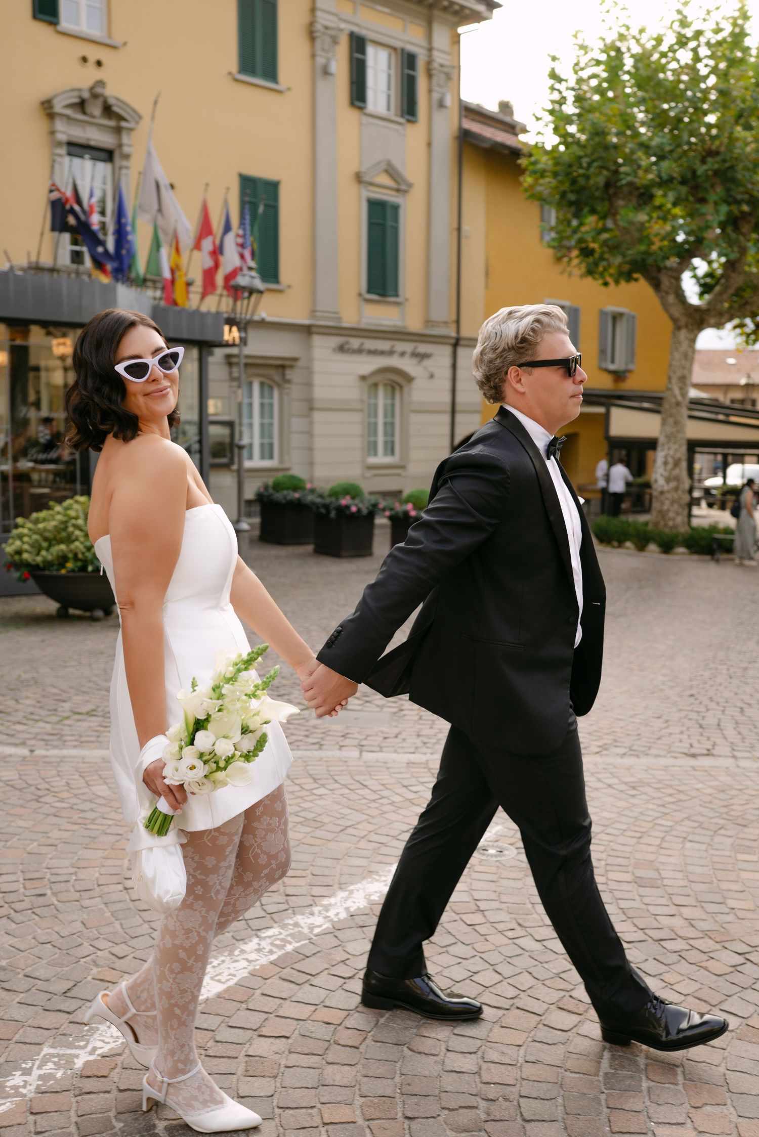 Bride in white mini dress and groom in black tuxedo holding hands walking through an Italian piazza.