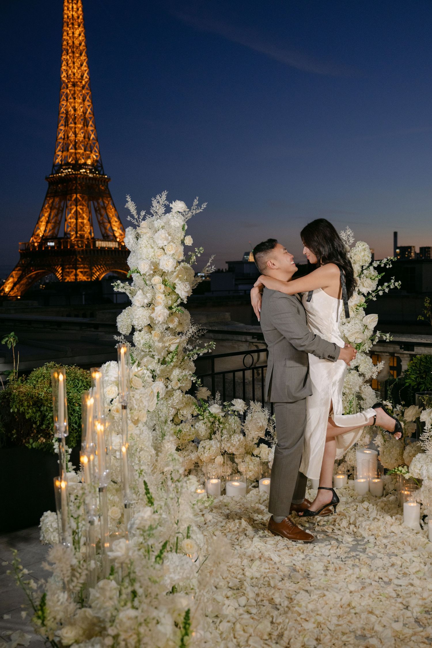 Couple embracing in a romantic Paris rooftop proposal setting with white flowers, candles, and the illuminated Eiffel Tower at dusk.