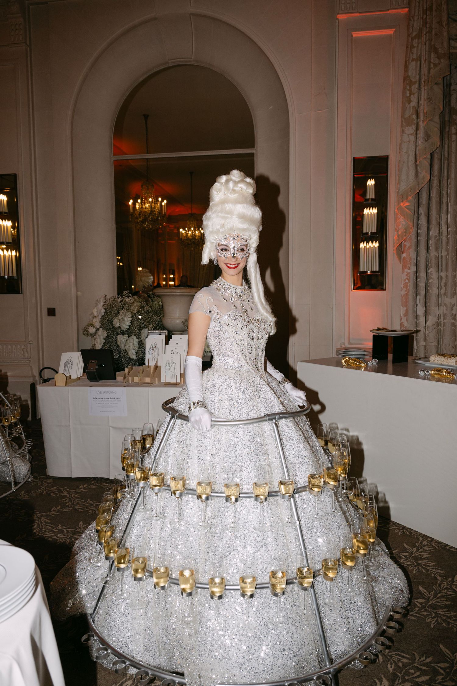 Champagne hostess in sparkling white gown with hoop skirt holding glasses tray at elegant event.